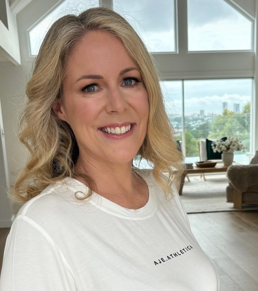A woman with blonde, shoulder-length curly hair and blue eyes smiling in a bright room with large windows and a cityscape view in the background.