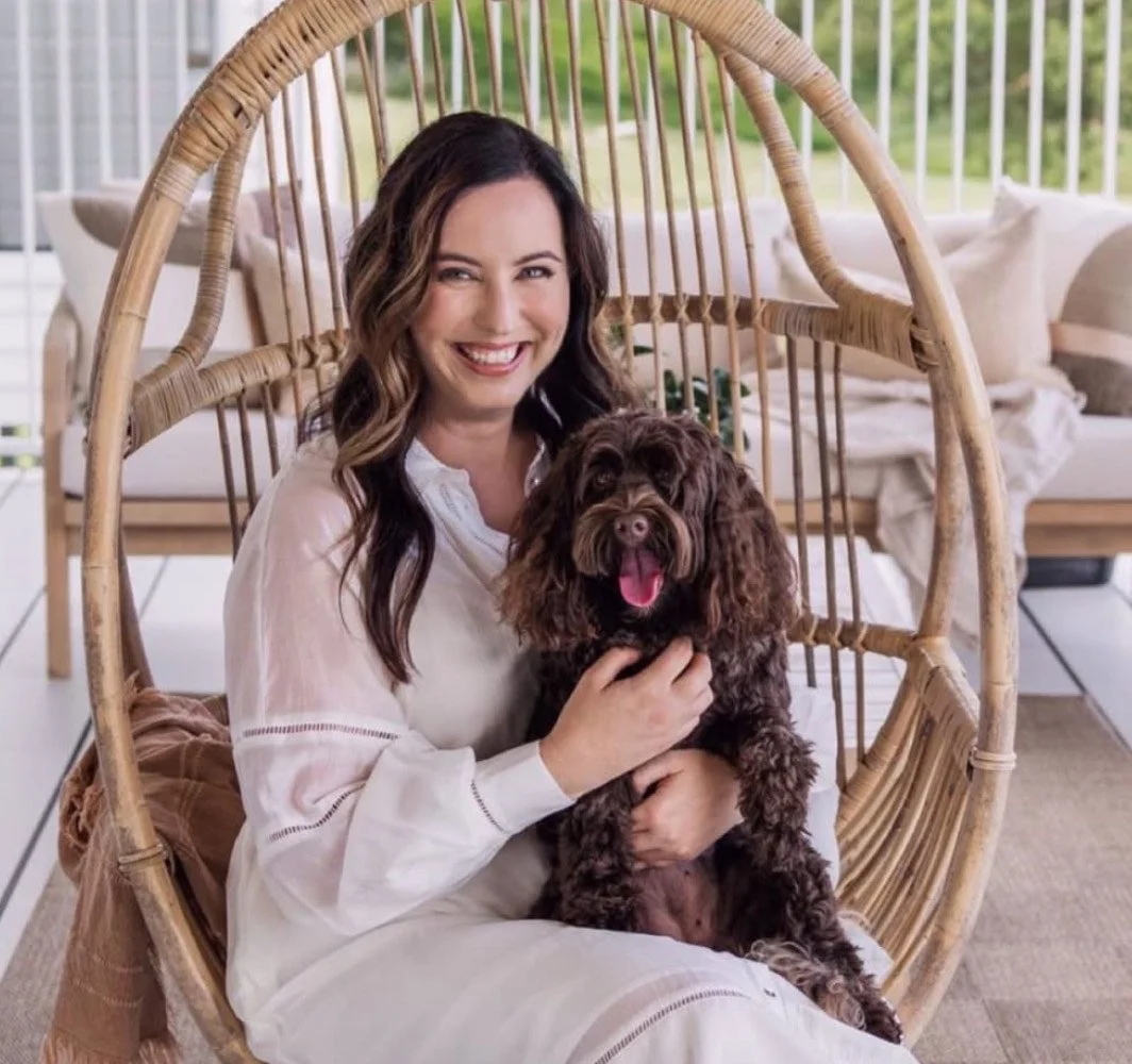 A woman with long brown hair smiling while sitting in a hanging wicker chair, holding a brown, curly-haired dog with its tongue out inside her lap. The background features an outdoor patio with cushions and a railing.