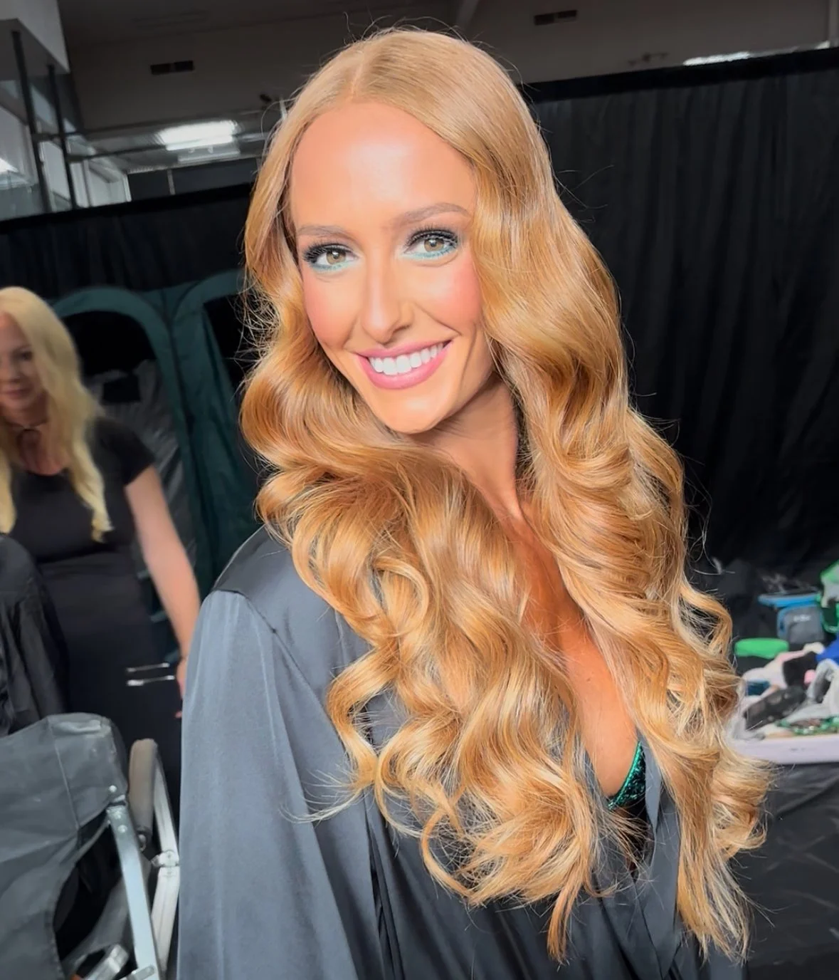 Close-up of a smiling woman with long wavy red hair, wearing a gray top, in a backstage or outdoor setting.