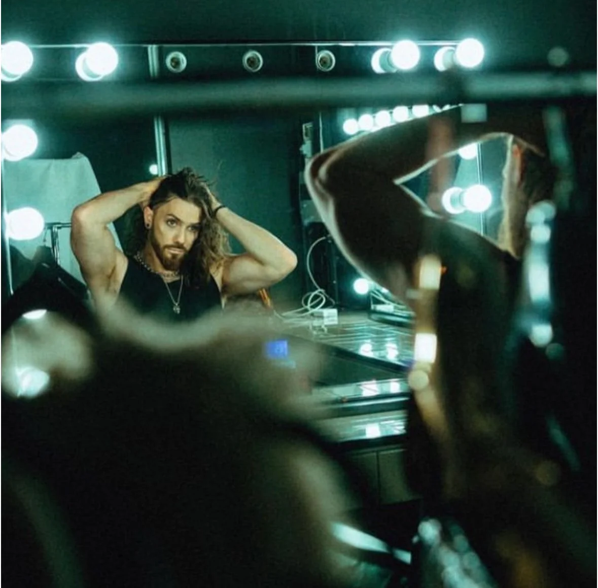 A man with shoulder-length curly hair and a beard, wearing a black sleeveless shirt, is looking at his reflection in a mirror and adjusting his hair in a dressing room with bright round lights around the mirror.