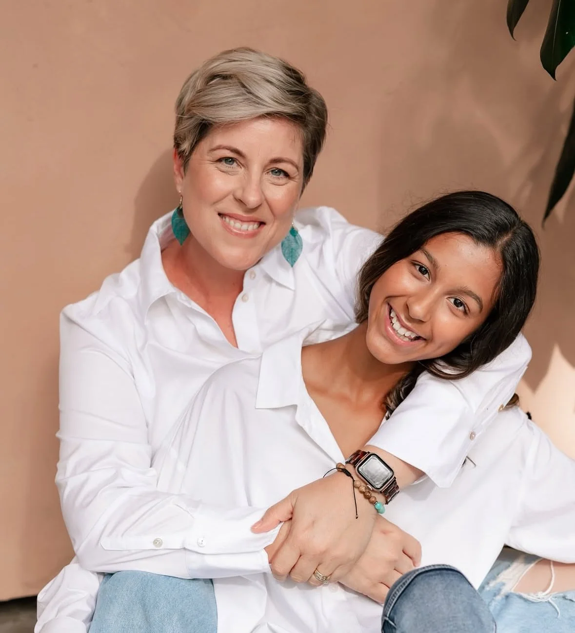 Two women smiling, one older with short blonde hair and the other younger with long dark hair, wearing white shirts, sitting closely together against a beige background.