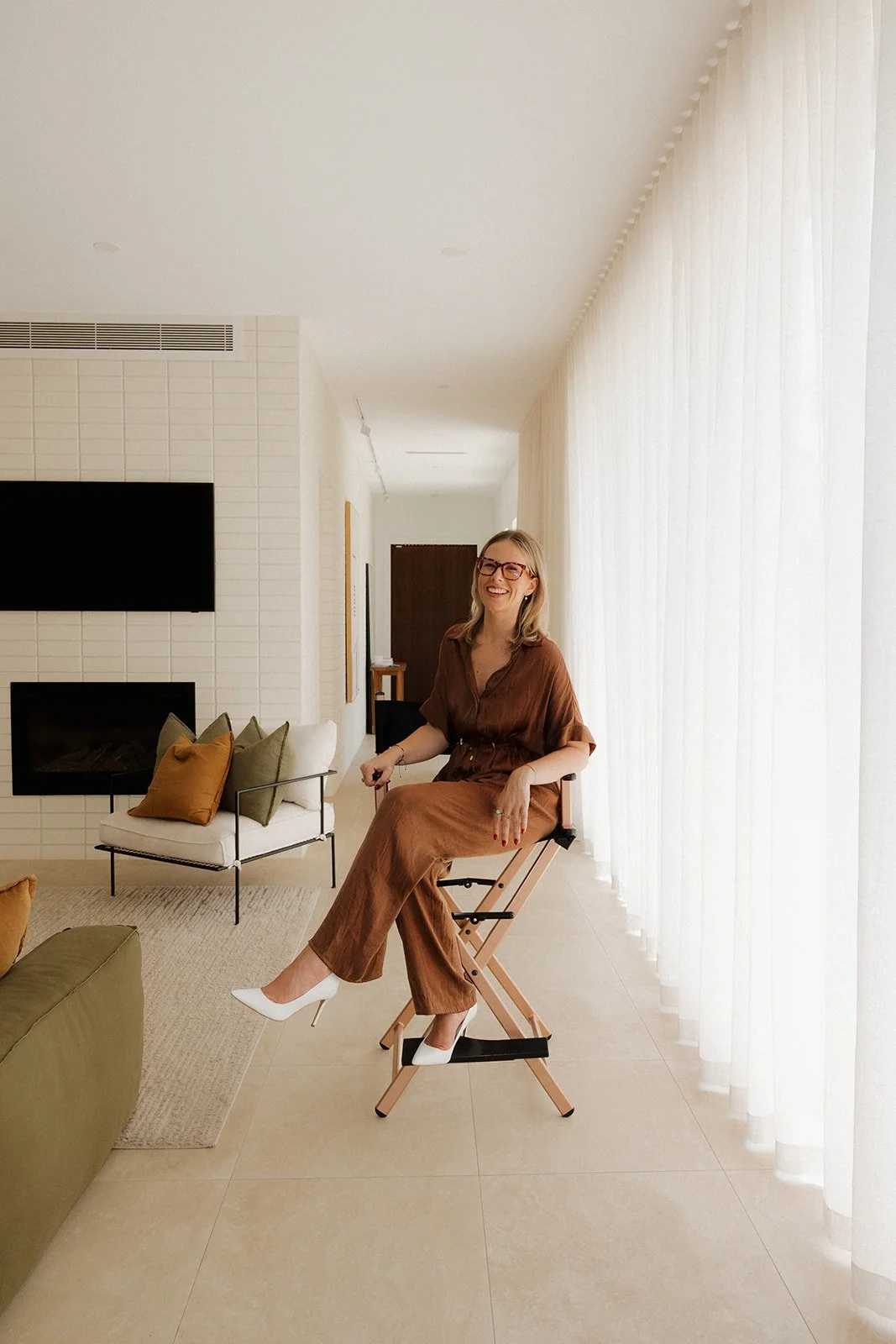 A woman sitting on a folding chair in a modern living room, smiling, wearing glasses, a brown outfit, and white high heels, with sunlight coming through sheer curtains on the right.