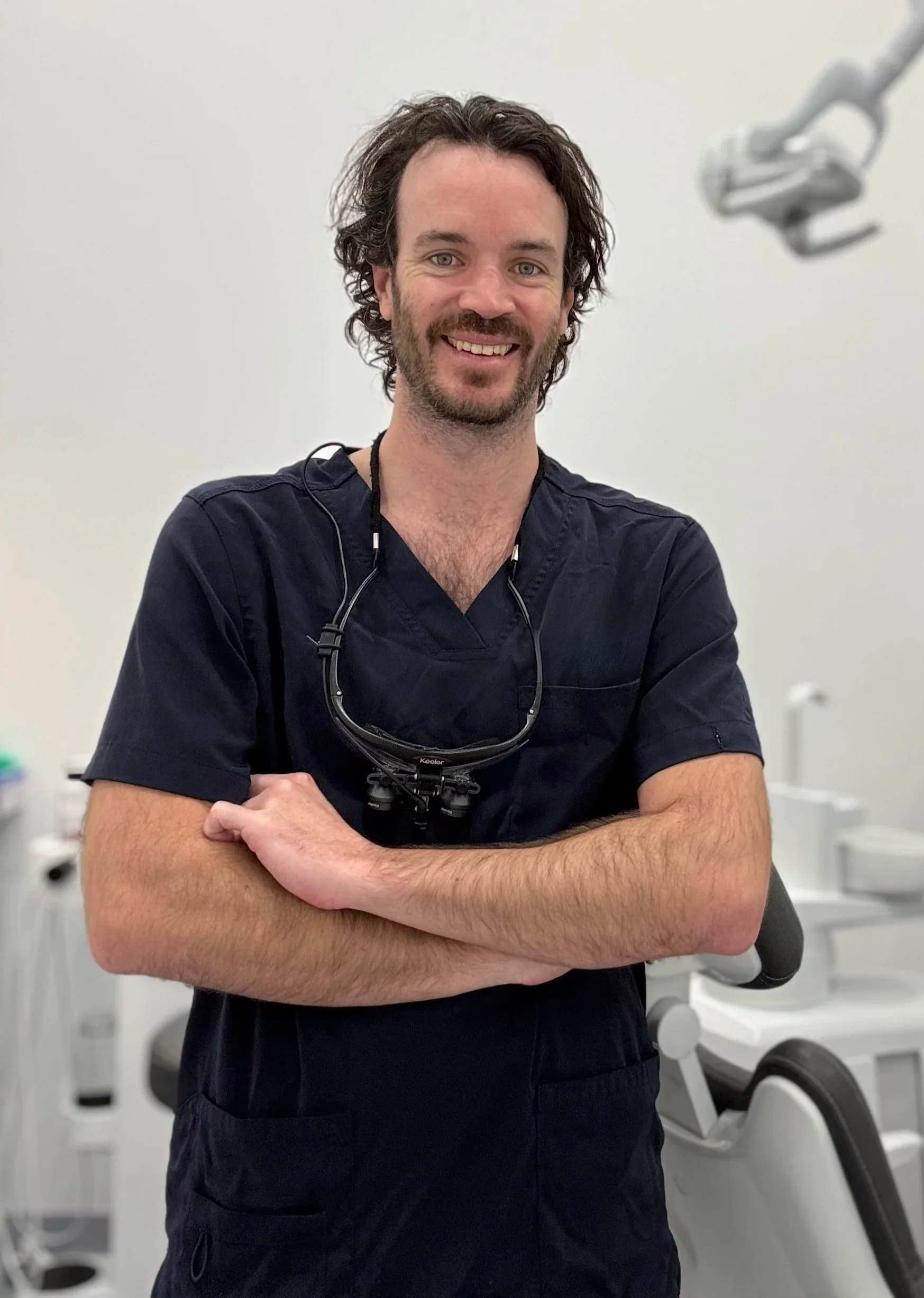 A male healthcare professional in navy scrubs stands in a medical room, smiling with arms crossed, wearing a pair of glasses around his neck.