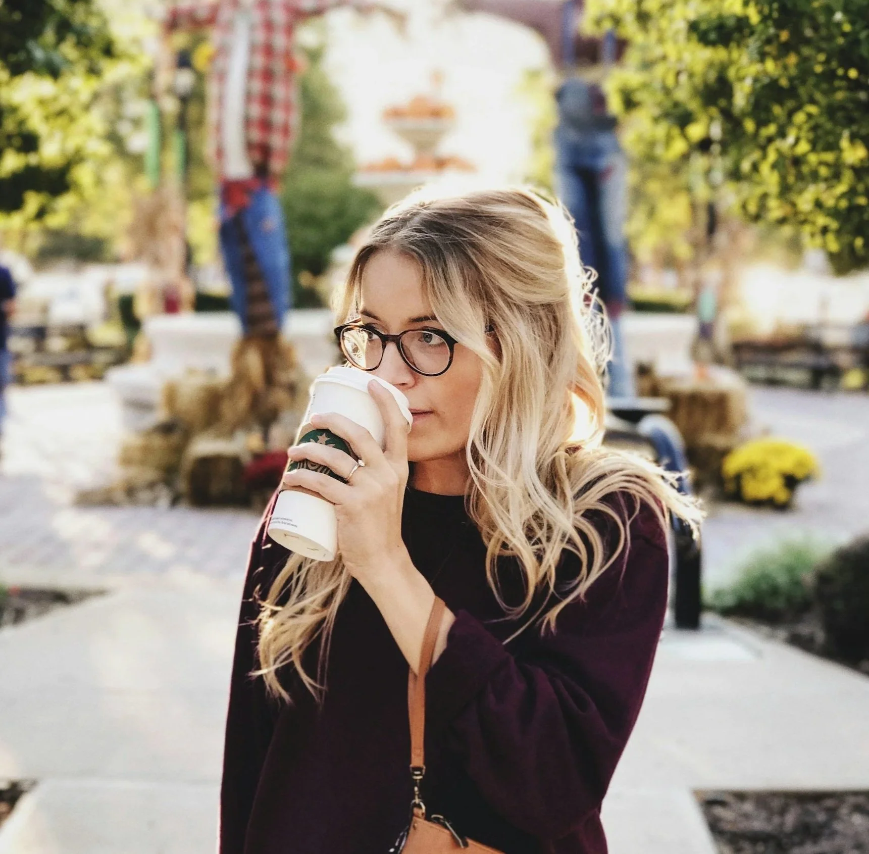 A woman with blonde wavy hair and glasses holding a Starbucks coffee cup, standing outdoors in a park-like setting with trees, benches, and blurred people in the background. teeth staining, teeth whitening