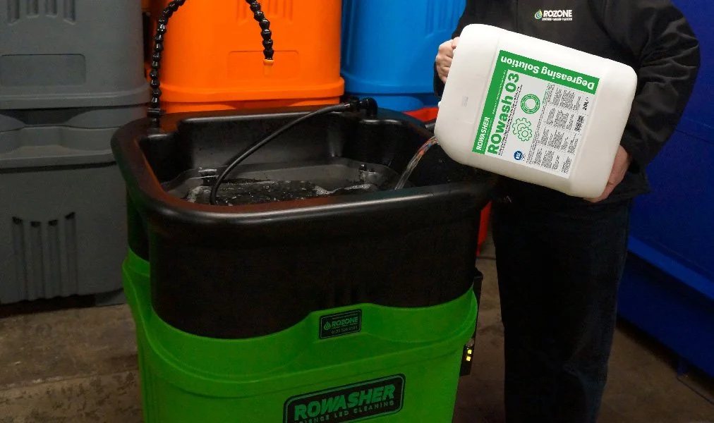 Person pouring ROwasher solution into a green ROwasher industrial cleaning machine, with gray, orange, and blue containers in the background.