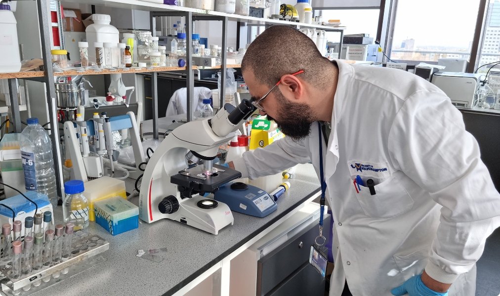 A scientist observing a sample through a microscope in a laboratory filled with lab equipment and supplies.
