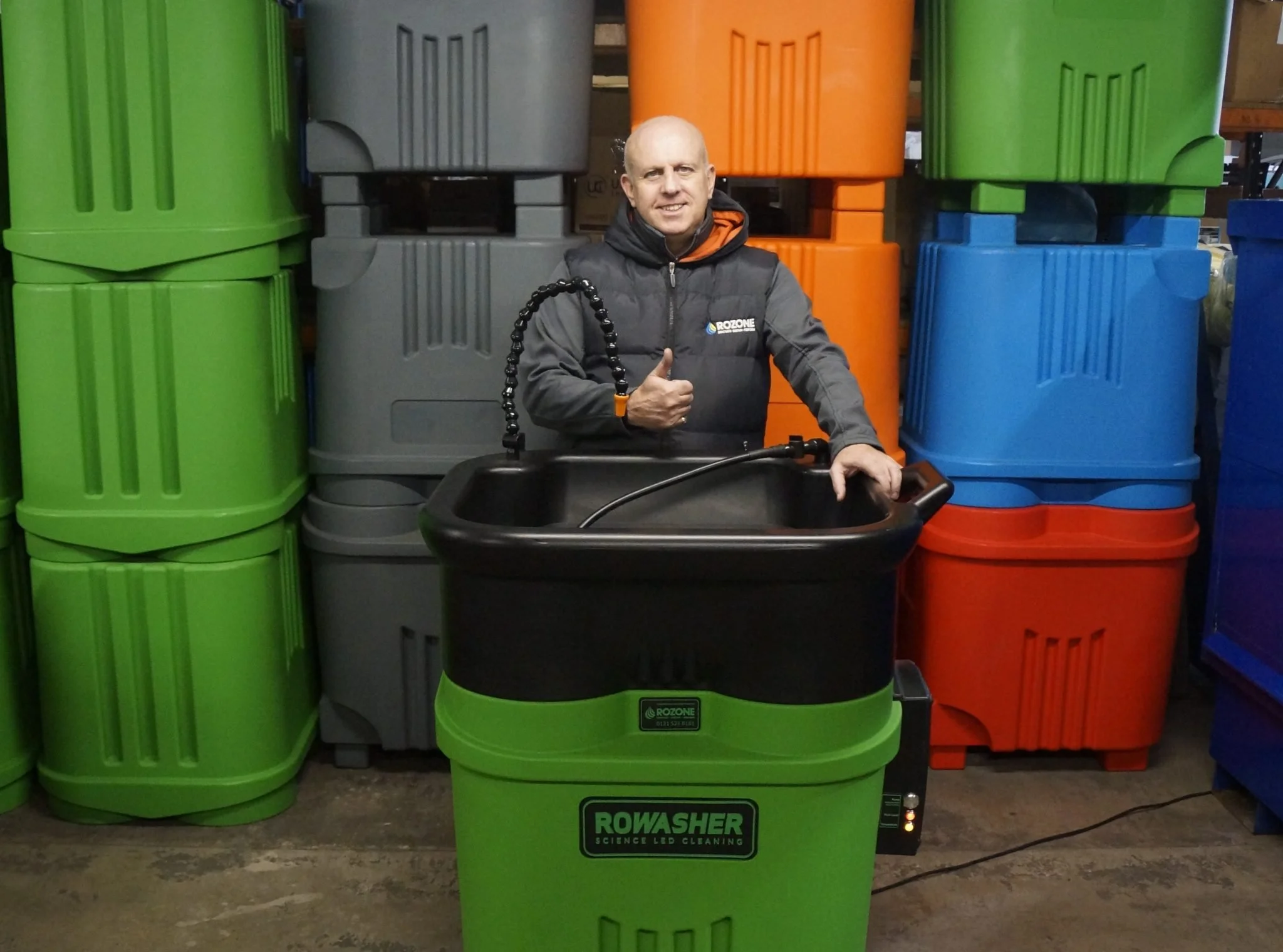 A man standing behind a green and black portable cleaning machine labeled 'ROWASHER' with a black tub and hose, giving a thumbs-up, in front of stacked colorful plastic storage bins.