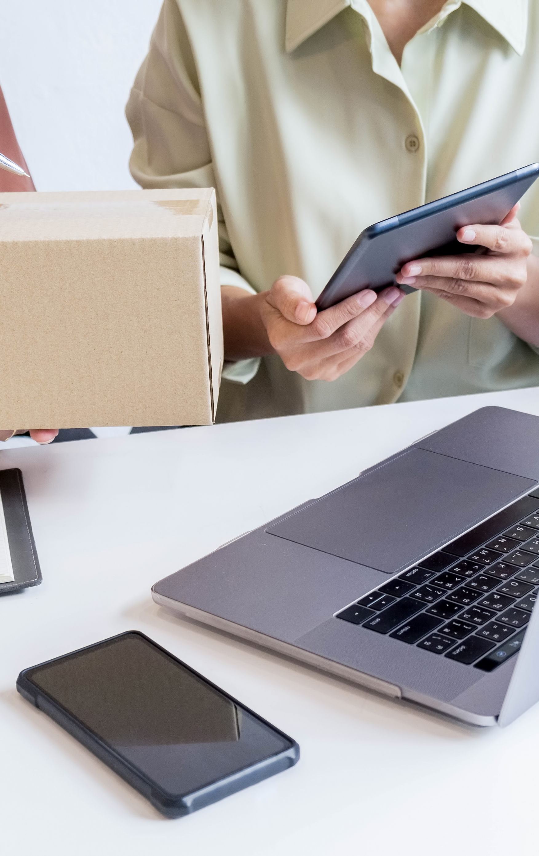 Person holding a cardboard box in one hand and a smartphone in the other, standing at a desk with a laptop, a smartphone, and a notebook.