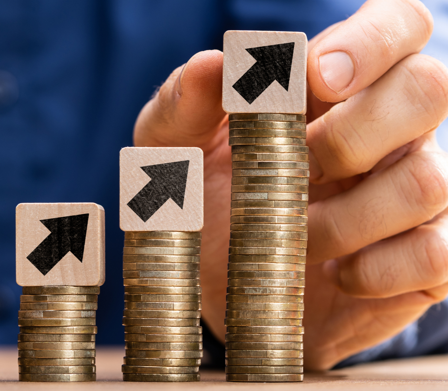 Stacks of coins increasing in height from left to right with wooden blocks bearing upward arrows on top of each stack being held in a hand, symbolizing growth or progress.