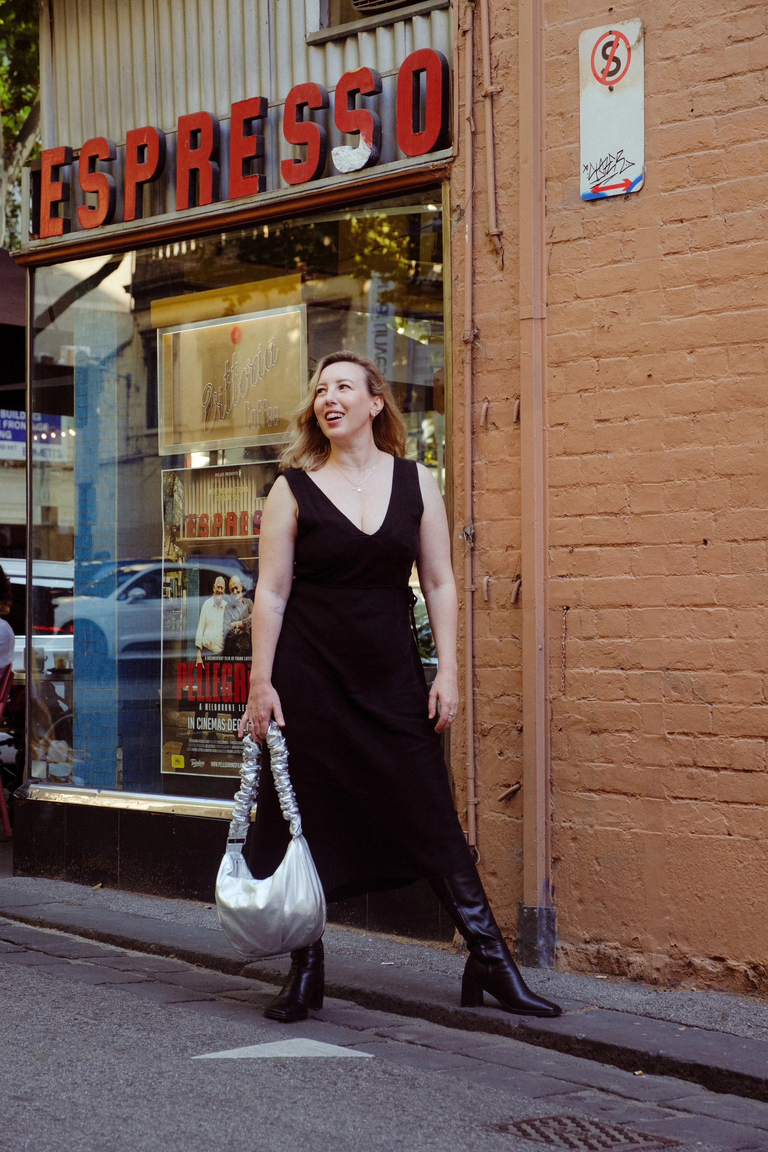 A woman in black dress and boots holding a silver bag, standing on a city sidewalk in front of a coffee shop with a sign that reads 'Espresso'.