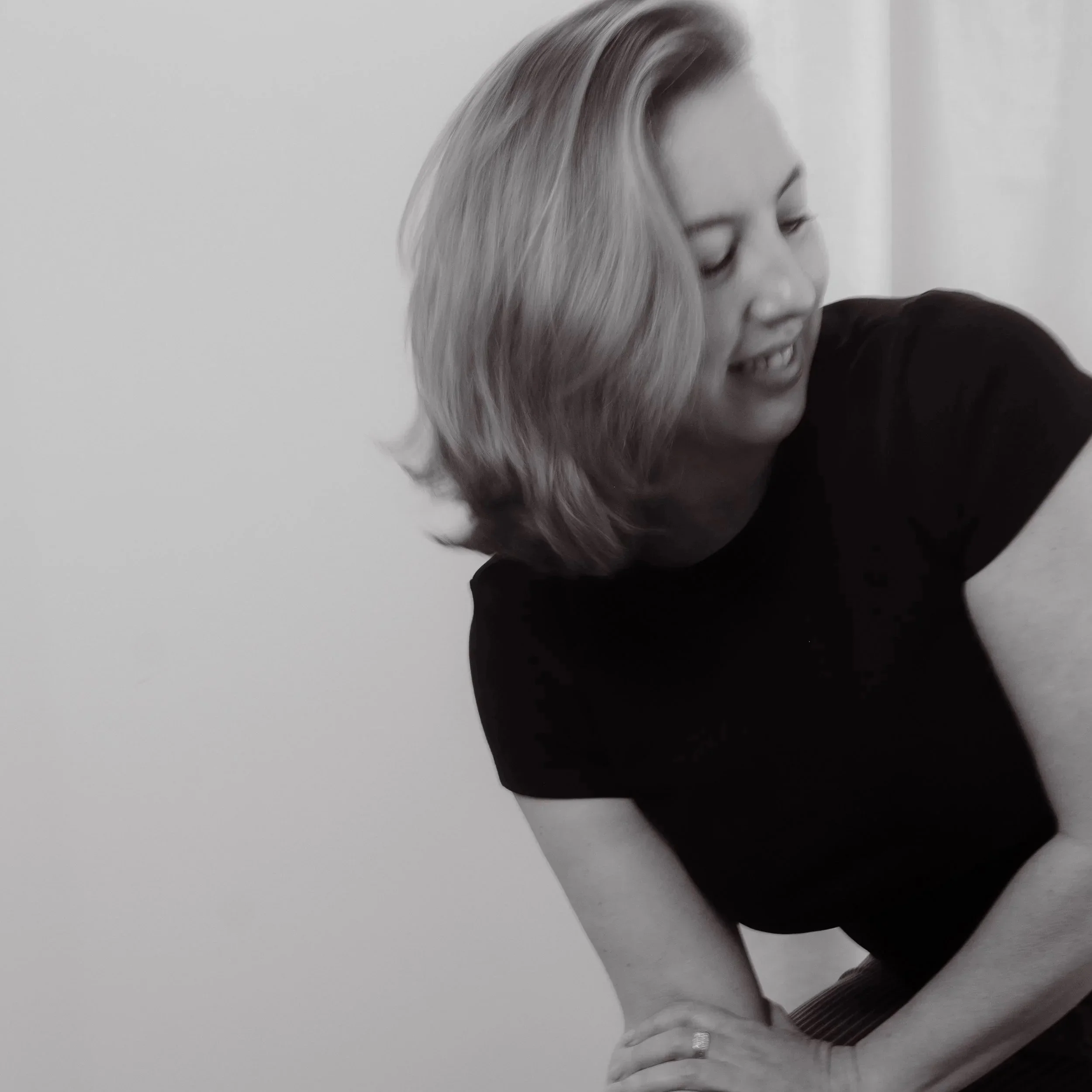 A black-and-white photo of a woman with shoulder-length hair, wearing a black shirt, smiling and looking down, with her hands resting on her lap.
