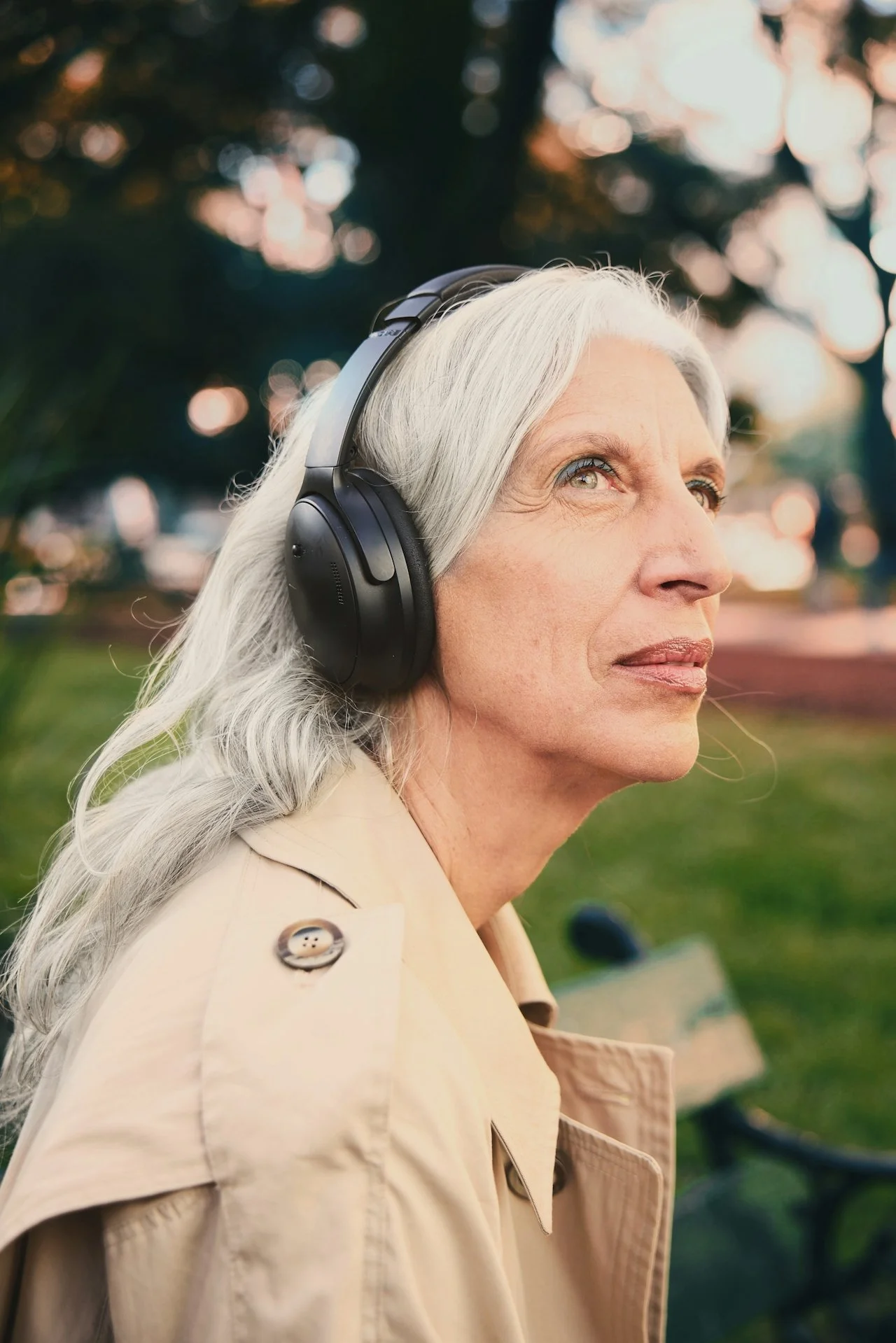 An older woman with long gray hair wearing a beige trench coat and black headphones outdoors during sunset, with trees and a park in the background.