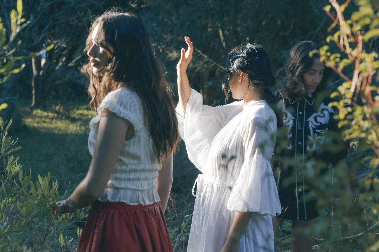 Three women standing outdoors among greenery during daytime, appearing to be in a serene or contemplative moment.