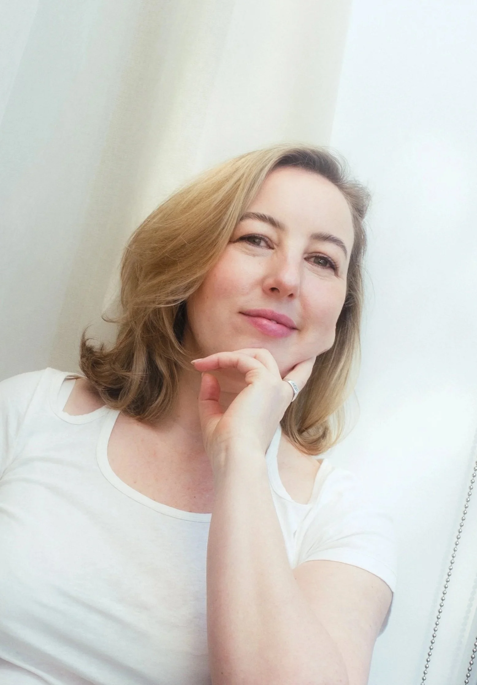 A woman with shoulder-length blonde hair, wearing a white top, is smiling softly and resting her chin on her hand, looking at the camera against a light-colored wall.