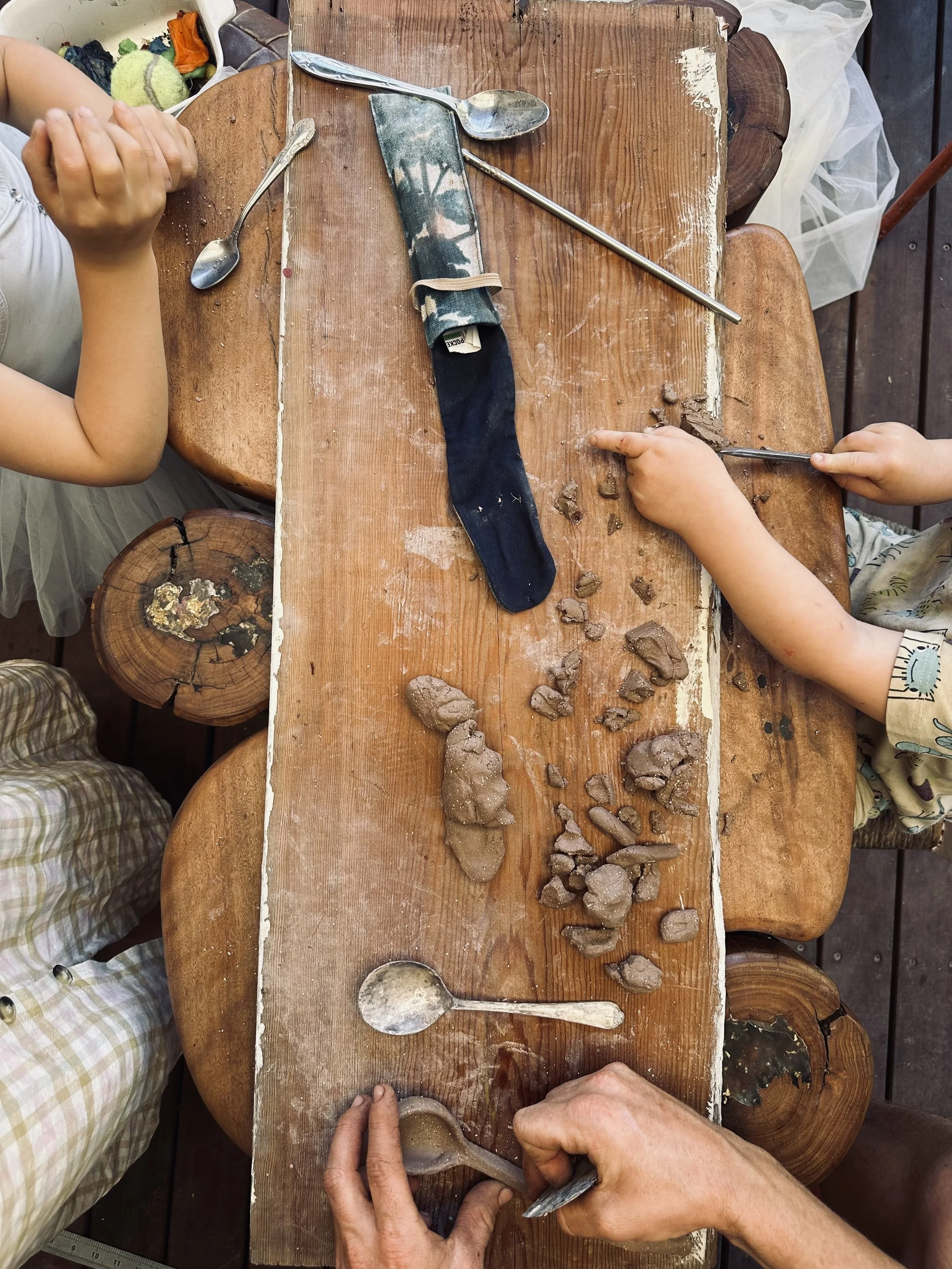 Children and adult at a wooden table dealing with clay or mud, with clay pieces, spoons, and a sock on the table.