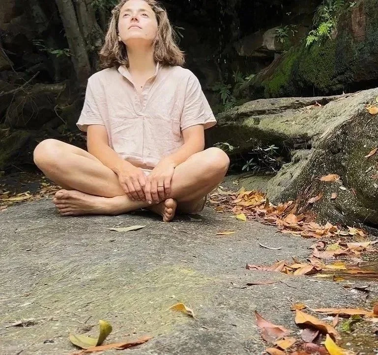A woman sitting cross-legged outdoors on a rock, with her hands resting on her feet, looking up, with greenery and large rocks in the background, and fallen leaves on the ground.