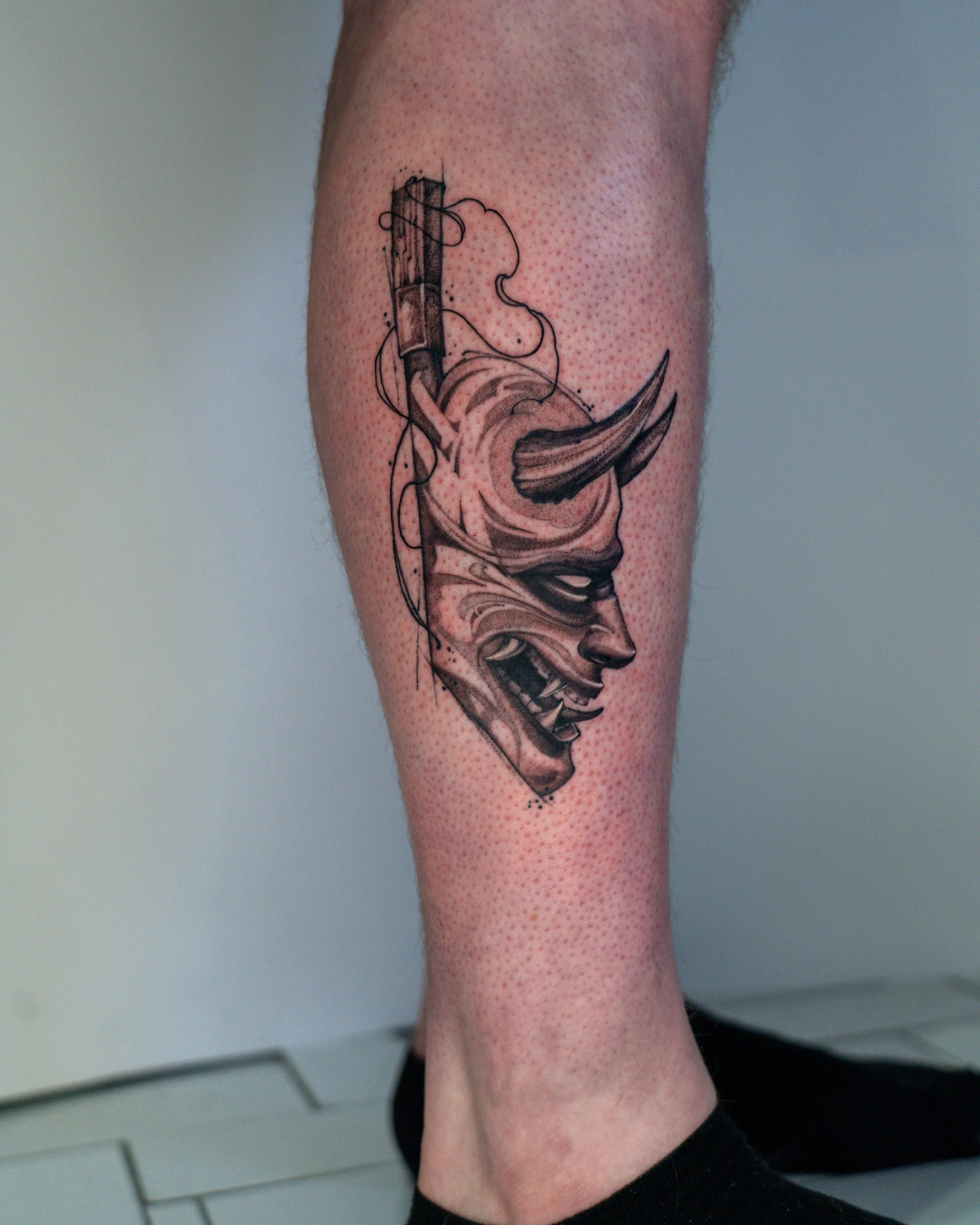Tattoo of an oni mask with sharp teeth, exaggerated features, and a jagged line on the forehead. The tattoo is done in black ink with detailed shading created by jay c tattoo