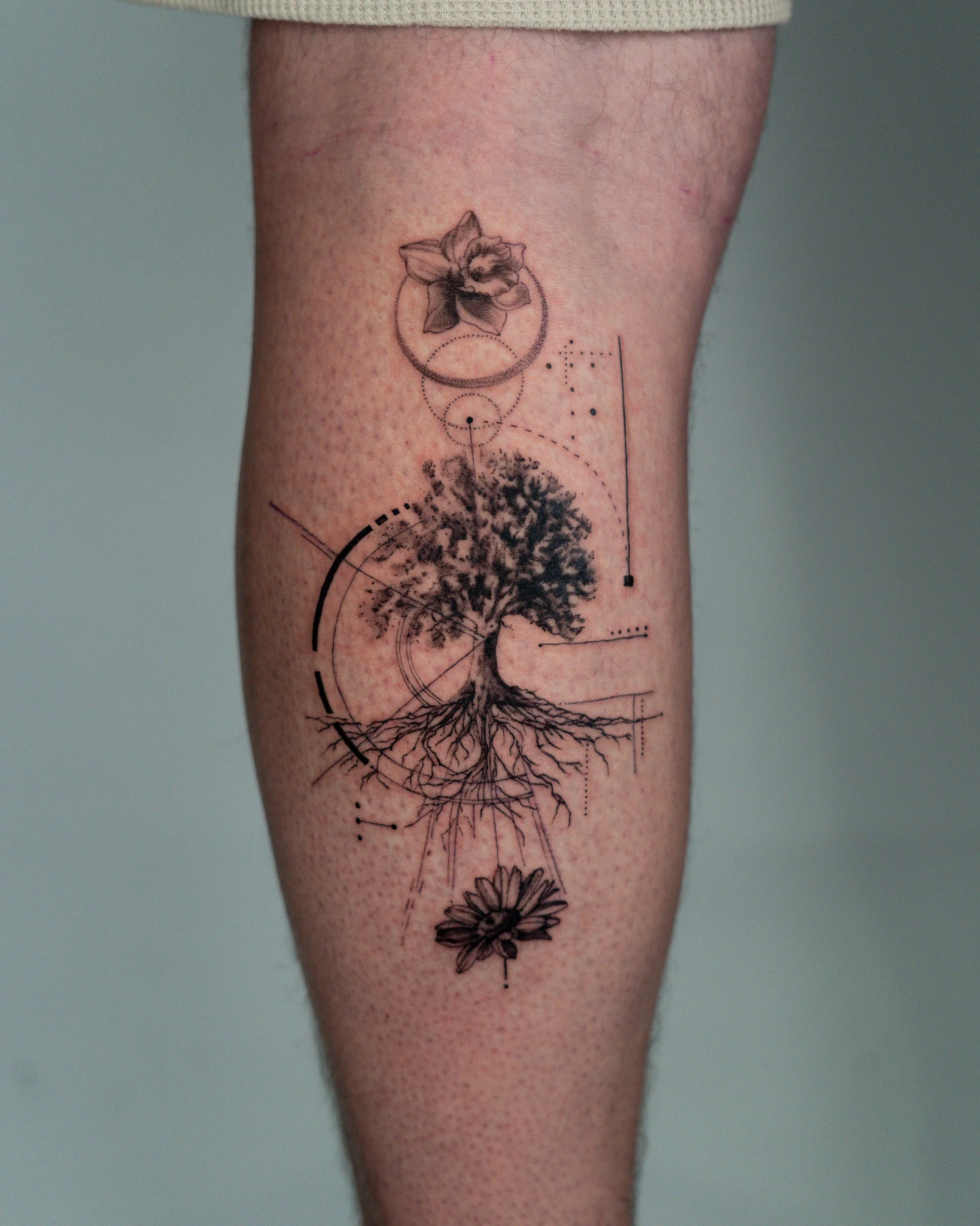 Fine line tattoo of a tree, geometric lines and dots, tree with roots, and a flower on a person's calf. created by jay c tattoo in brighton.