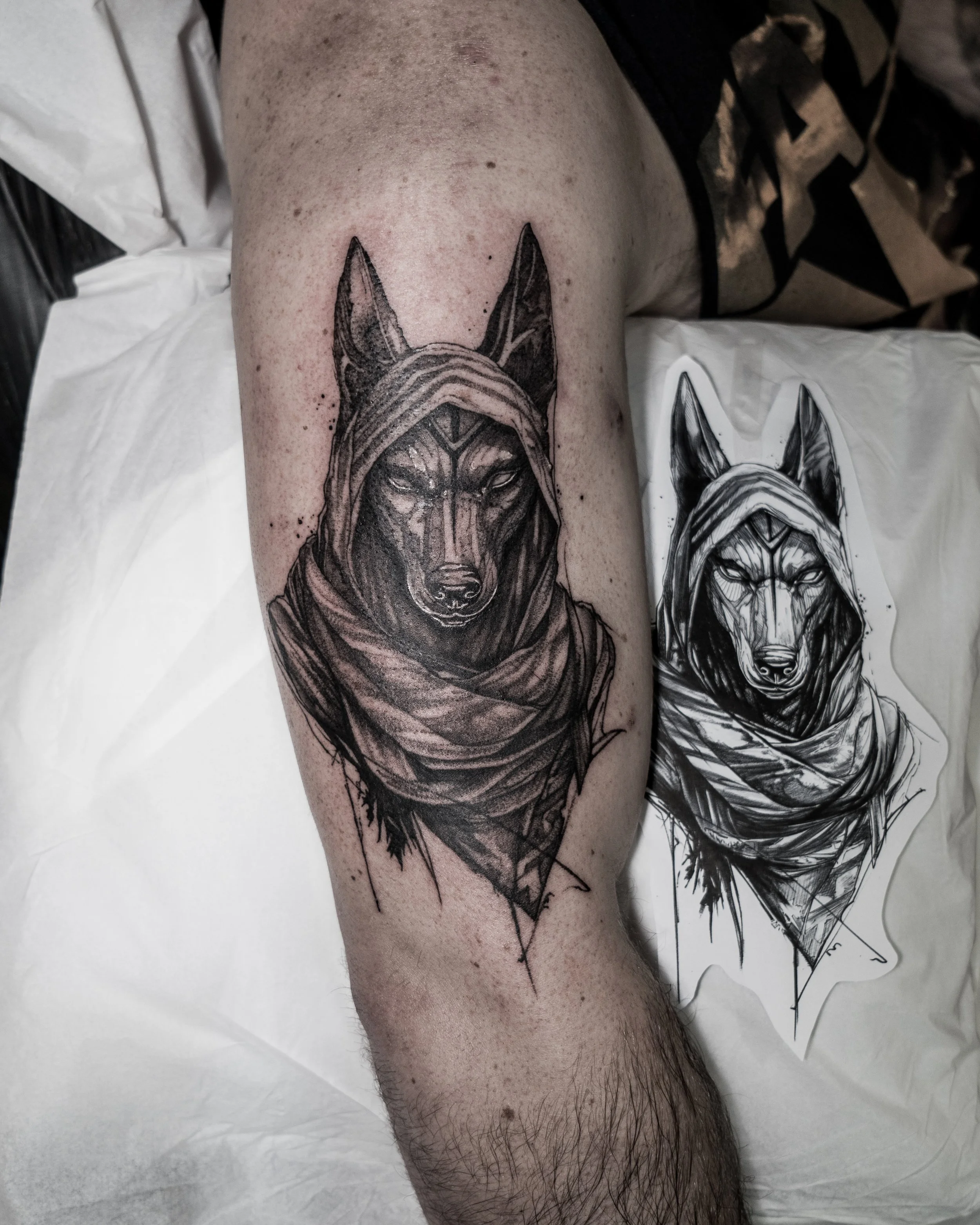 Black and grey tattoo of a Anubis wearing a hooded cloak on upper arm with matching sketch by Jay C Tattoo, Brighton and Hove tattoo artist, UK
