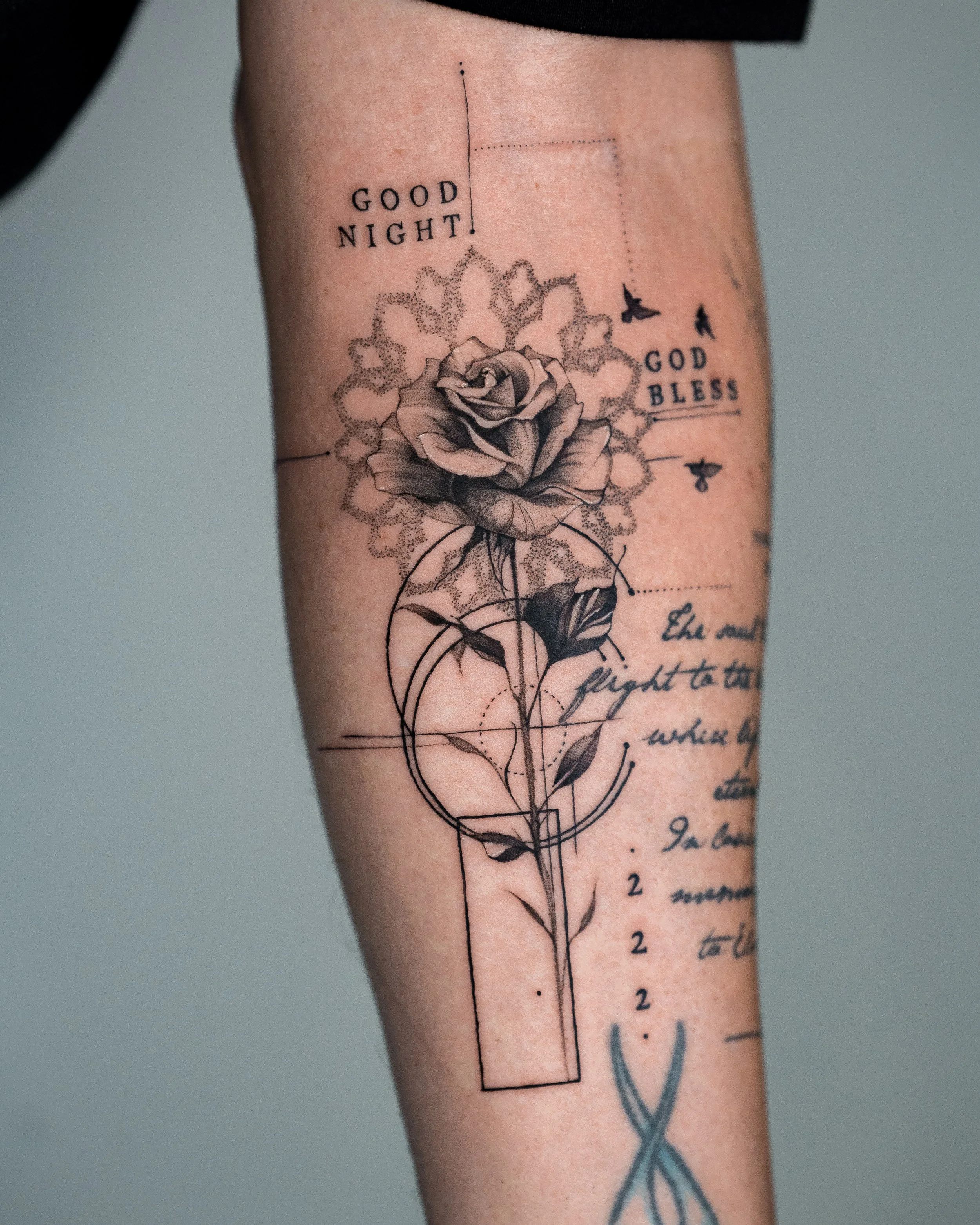 Tattoo on a person's arm featuring a detailed rose, geometric shapes, text reading 'GOOD NIGHT!' and 'GOD BLESS,' small birds, and handwritten script.