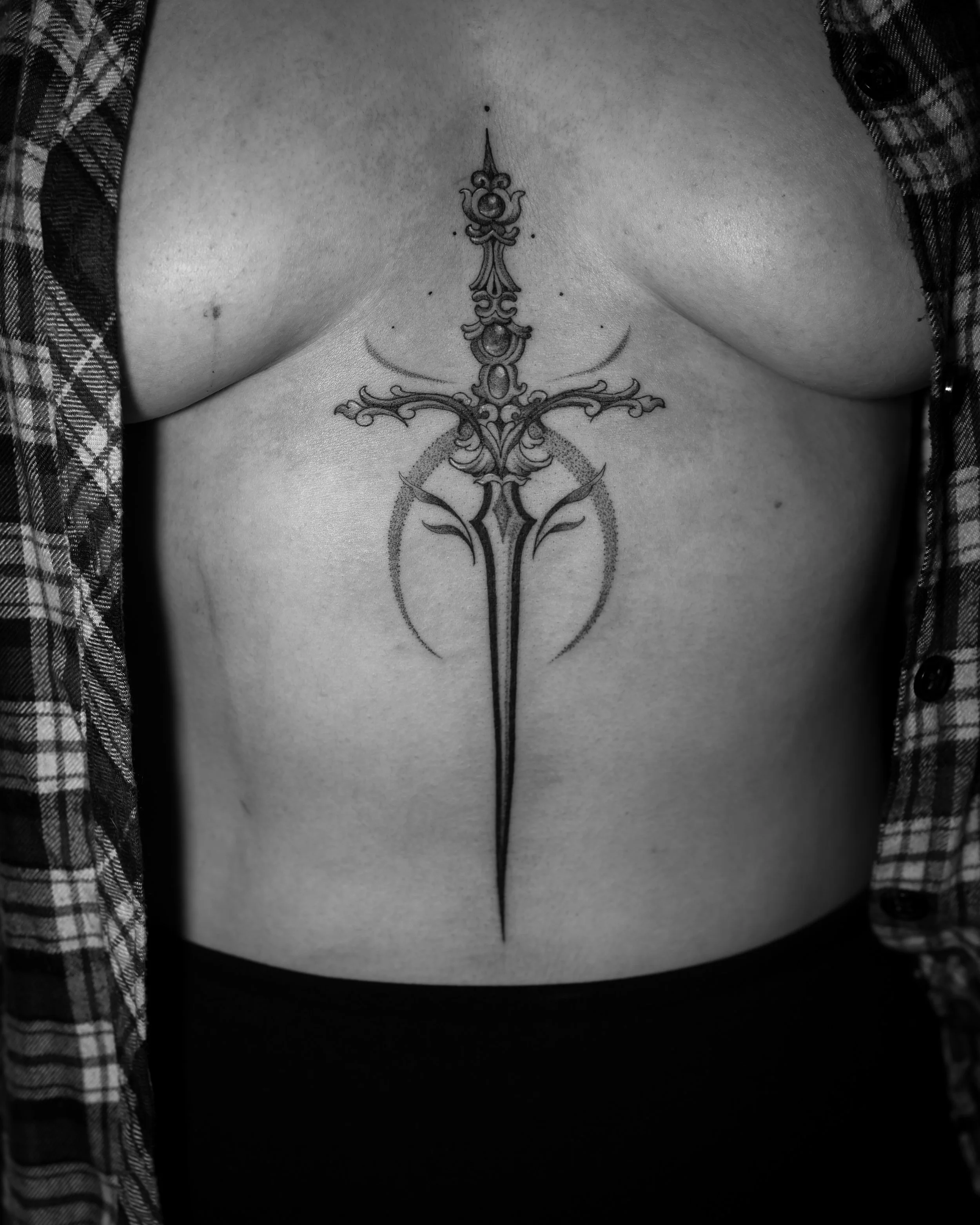 Black ink sternum tattoo of an ornate sword with symmetrical decorative elements by Jay C Tattoo, Brighton tattoo artist, UK
