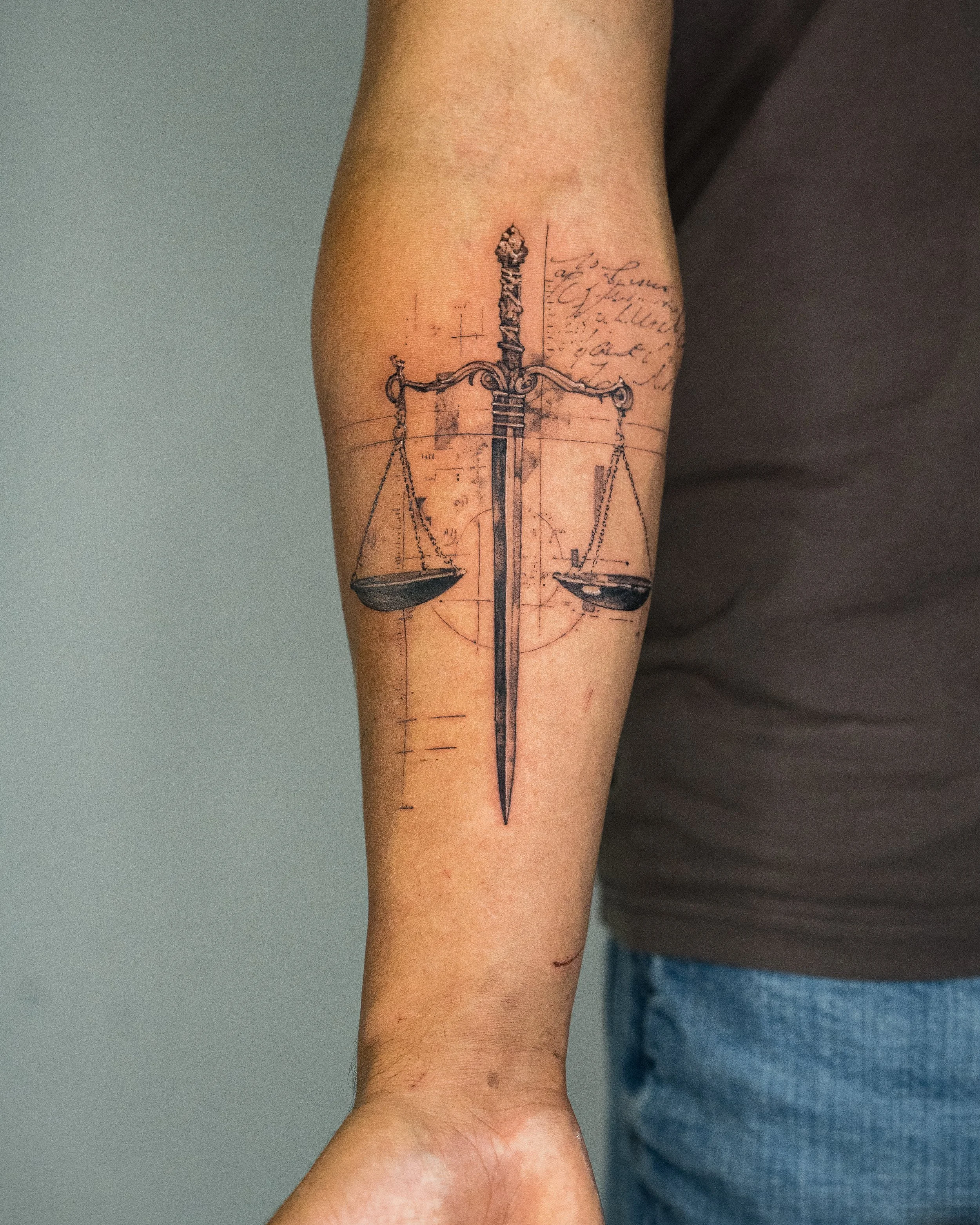 Tattoo of a balanced scale with a detailed sword in the center on a person's forearm, with sketches and handwritten notes in the background.
