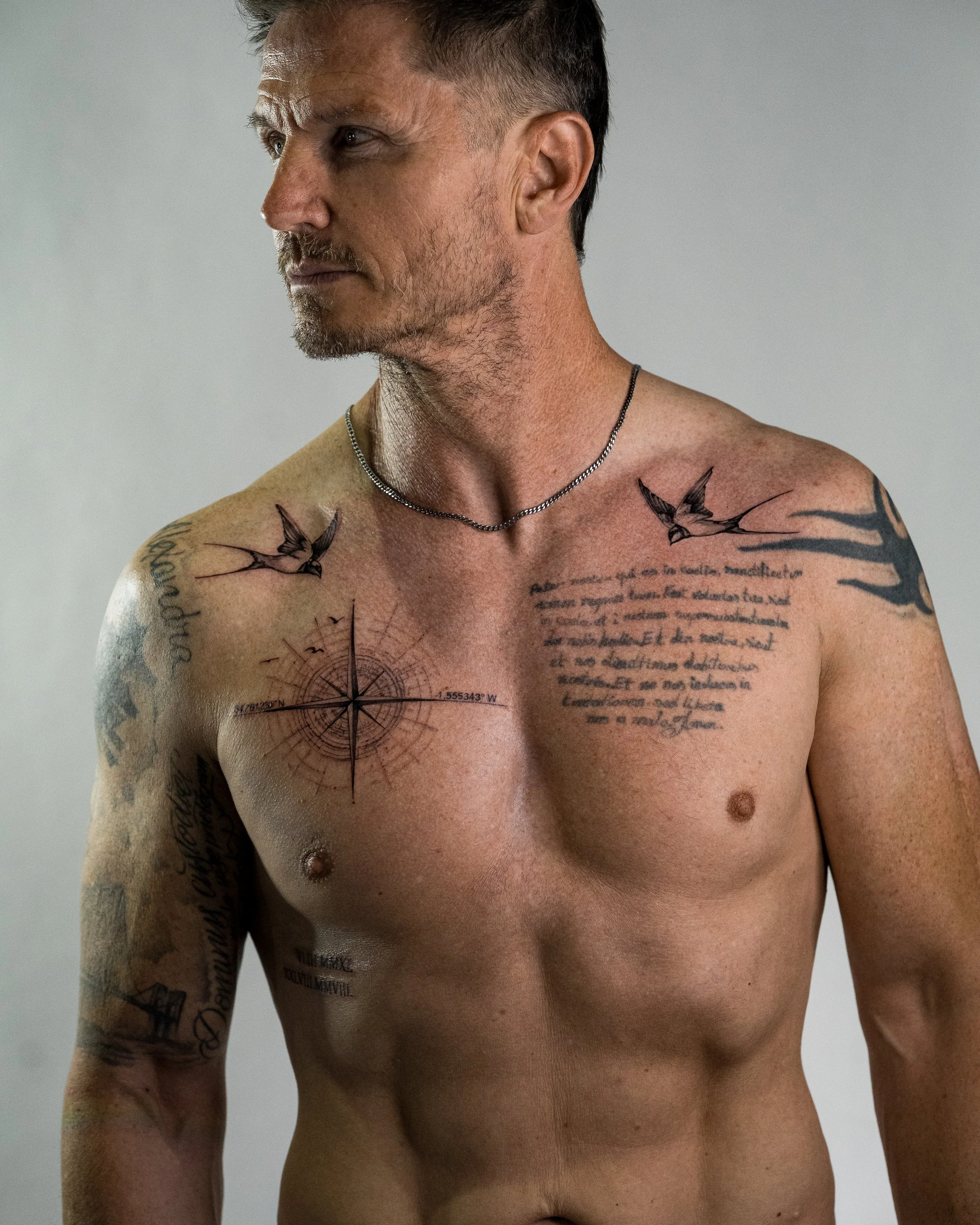 Close-up of a shirtless man with various tattoos on his chest, shoulders, and arms, wearing a silver chain necklace, with a neutral background.
