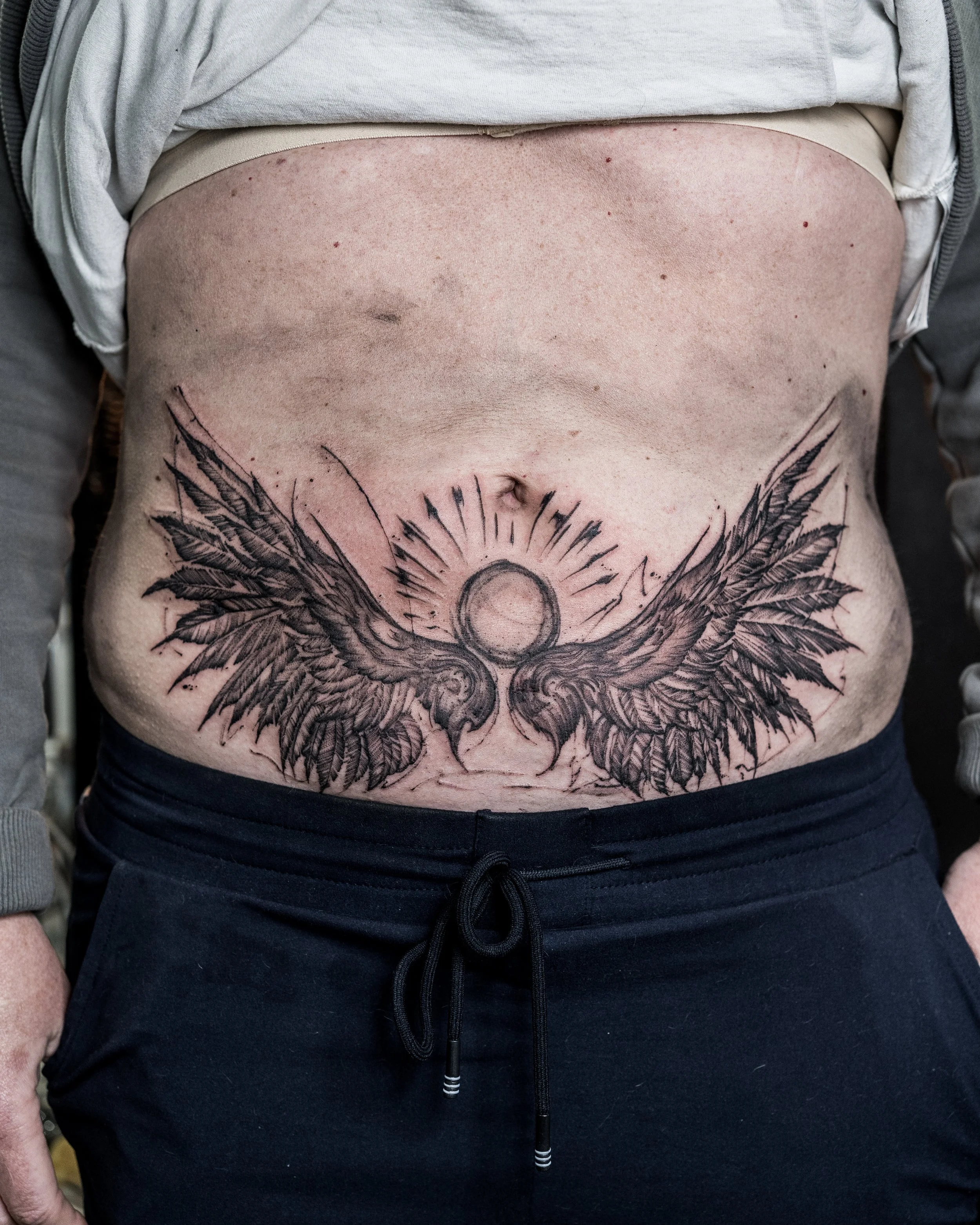 Large black ink lower abdomen tattoo of a sun with rays and wings by Jay C Tattoo, Brighton tattoo artist, UK
