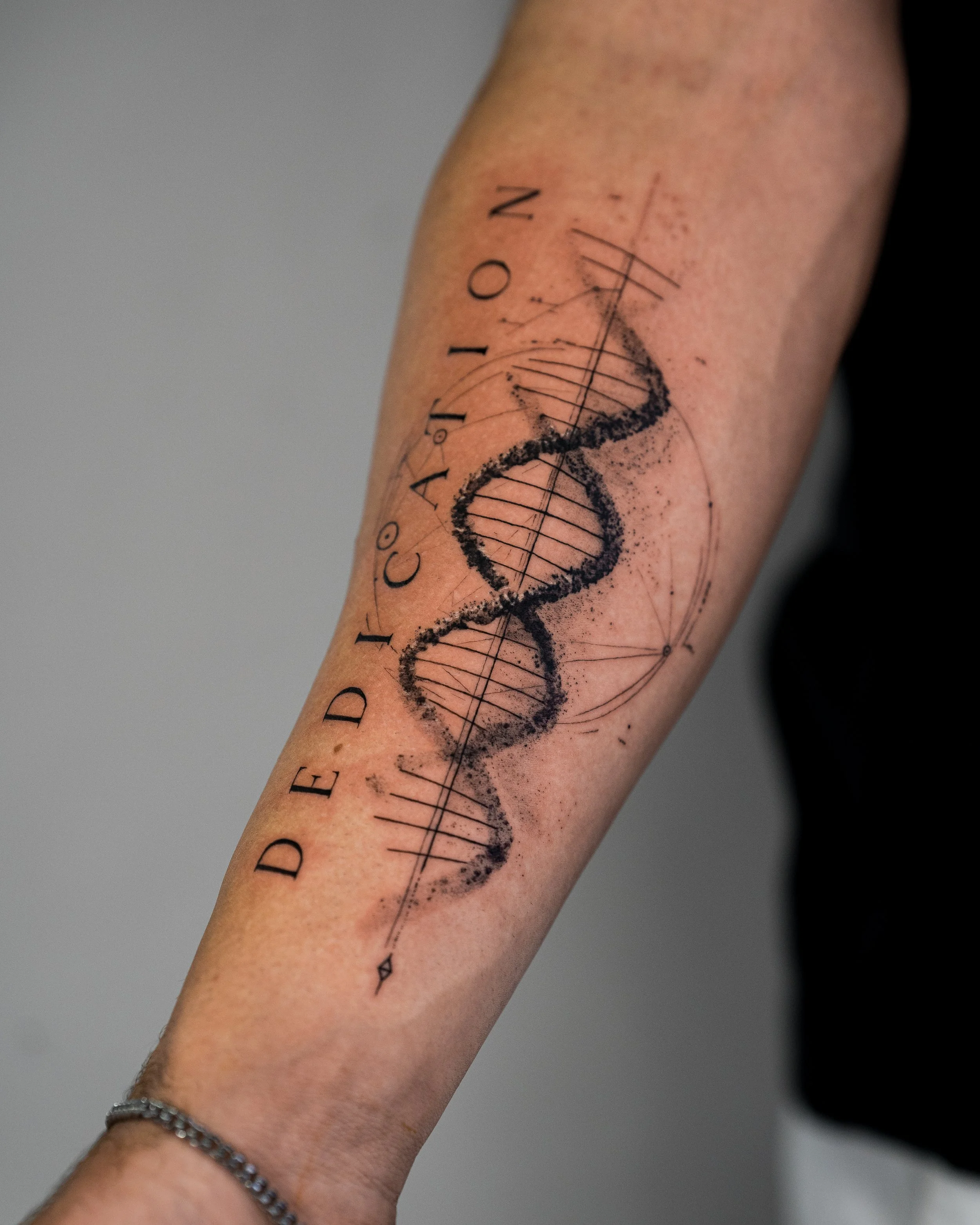 Close-up of a forearm with a tattoo of a mathematical geometric design, including a double helix DNA strand and anatomical text, with a silver chain bracelet.