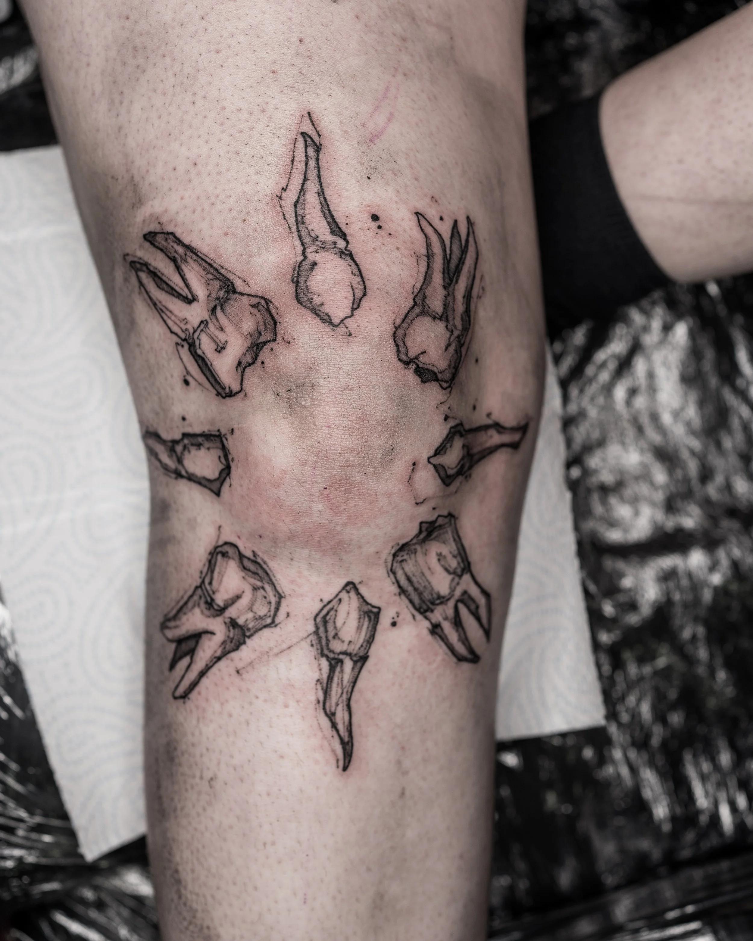 Black ink tattoo of broken teeth arranged in a circular pattern on a knee by Jay C Tattoo, Brighton and Hove tattoo artist, UK

