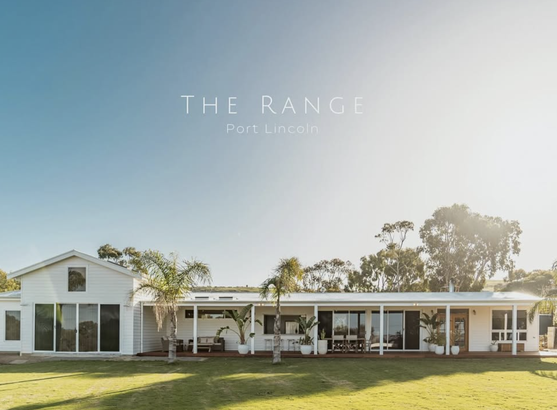 Modern white house with large glass doors and windows, surrounded by palm trees on a lush green lawn under a clear sky.