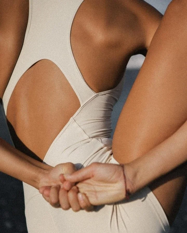Two women are holding hands, one wearing a beige dress with cutouts and the other wearing a black top.