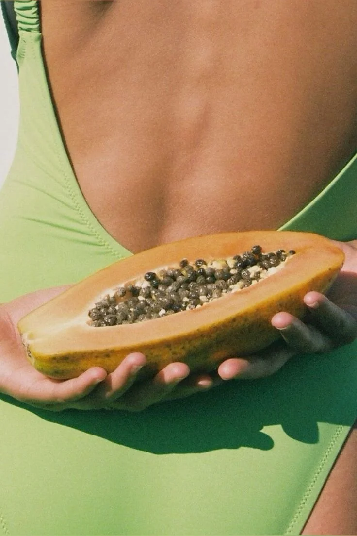 Close-up of a person holding a halved papaya fruit filled with black seeds, wearing a bright green top.