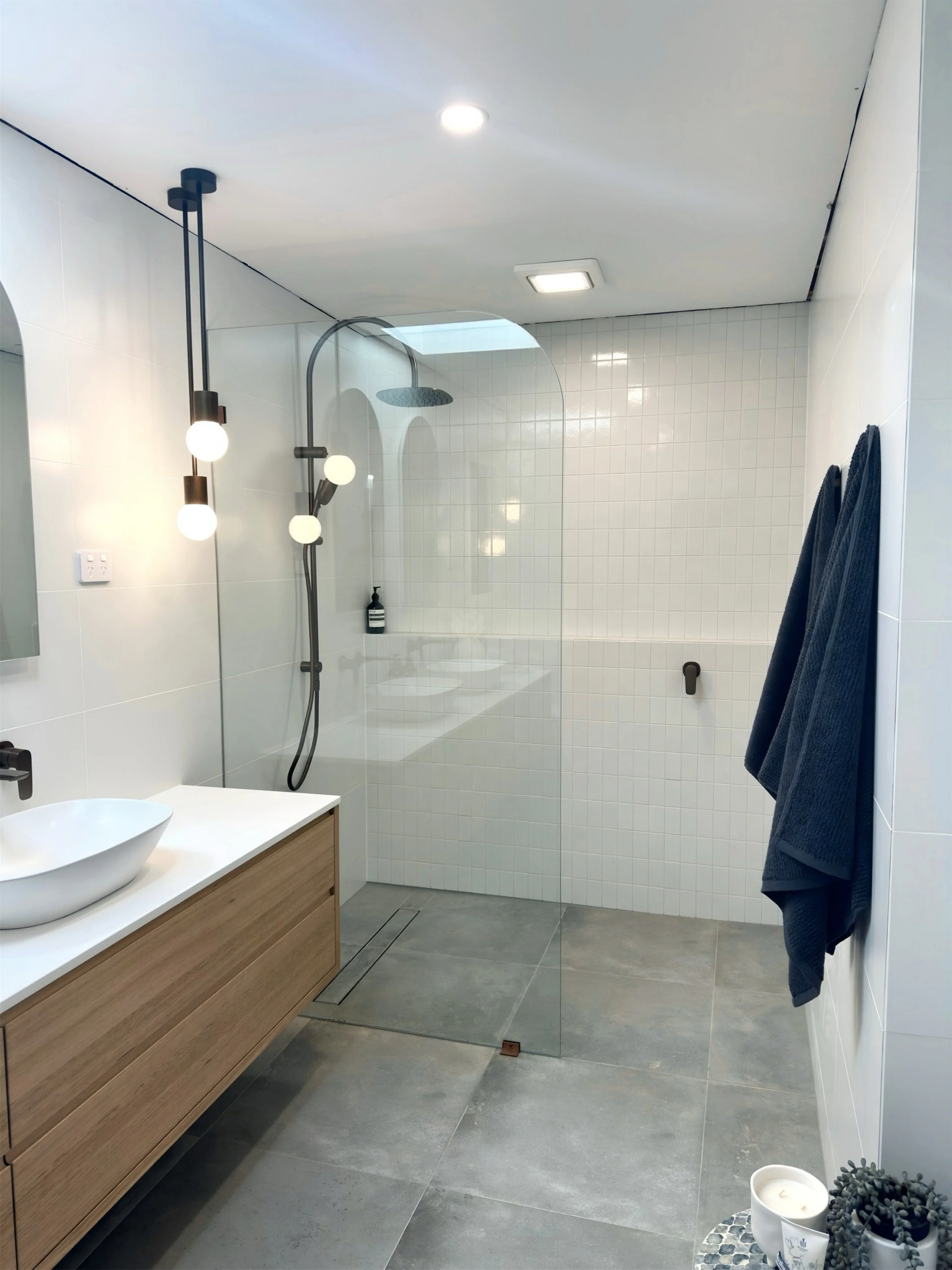 Modern bathroom with a walk-in shower, white tiled walls, gray floor tiles, a wooden vanity with a white vessel sink, pendant lighting, and dark towels hanging on the wall.
