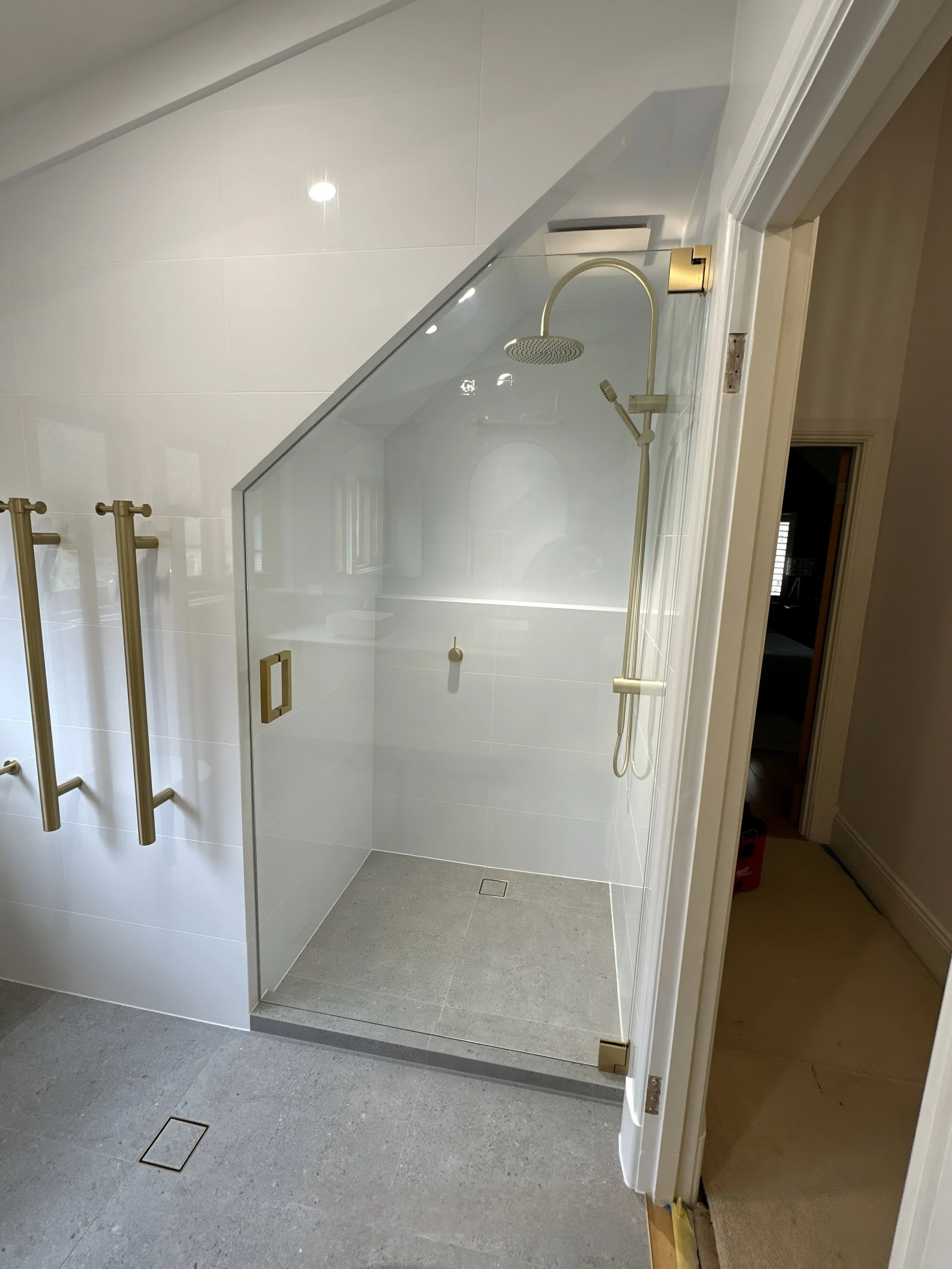 A modern bathroom with a walk-in shower enclosed by a glass wall. The shower has a ceiling-mounted showerhead and a handheld shower on a gold fixture. The floor is tiled with gray tiles, and the walls are white. There are metallic gold grab bars on the exterior wall of the shower.