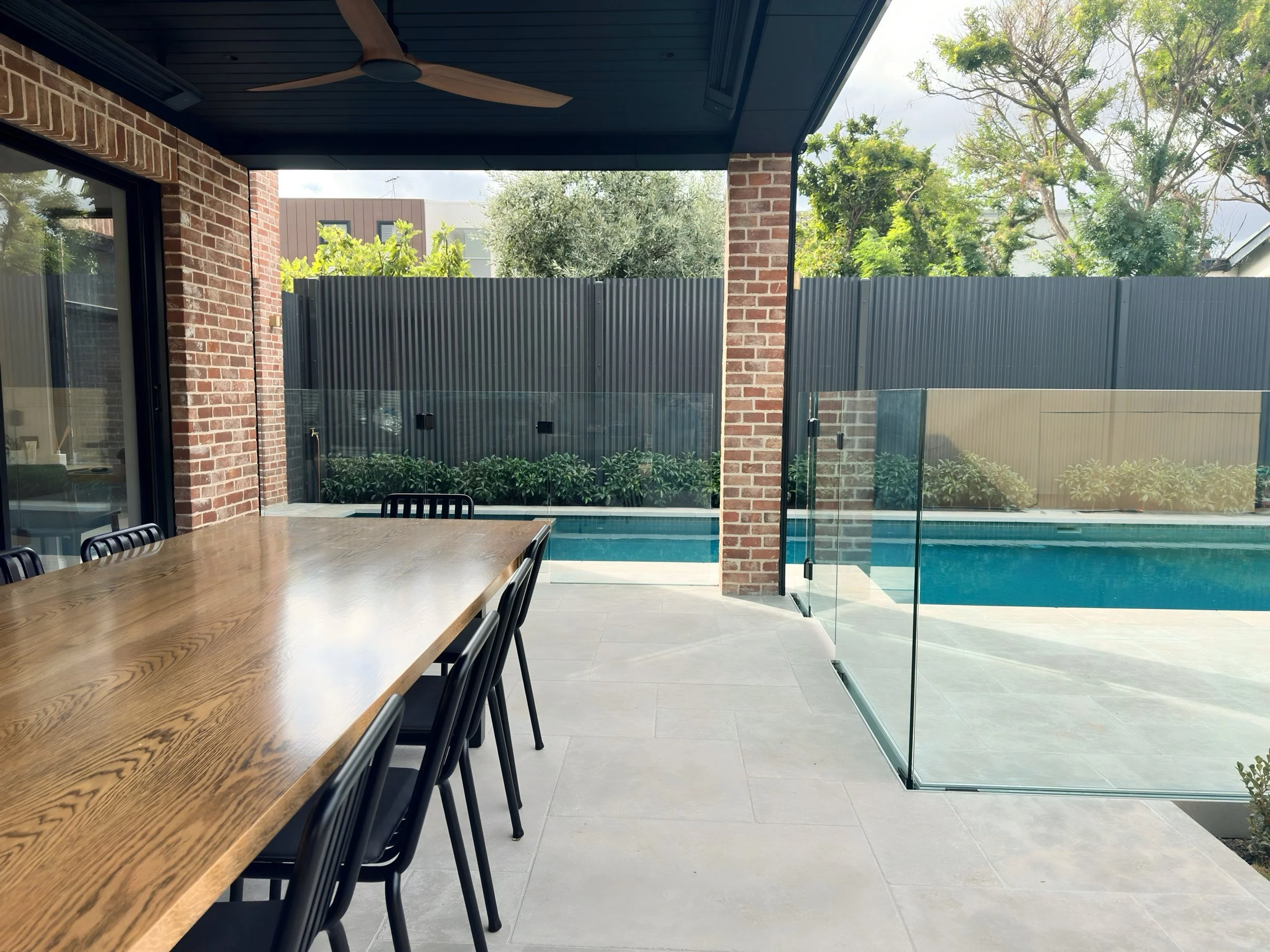 Outdoor patio with a wooden table and black chairs, overlooking a swimming pool enclosed by glass panels and a tall black fence, surrounded by trees and plants.