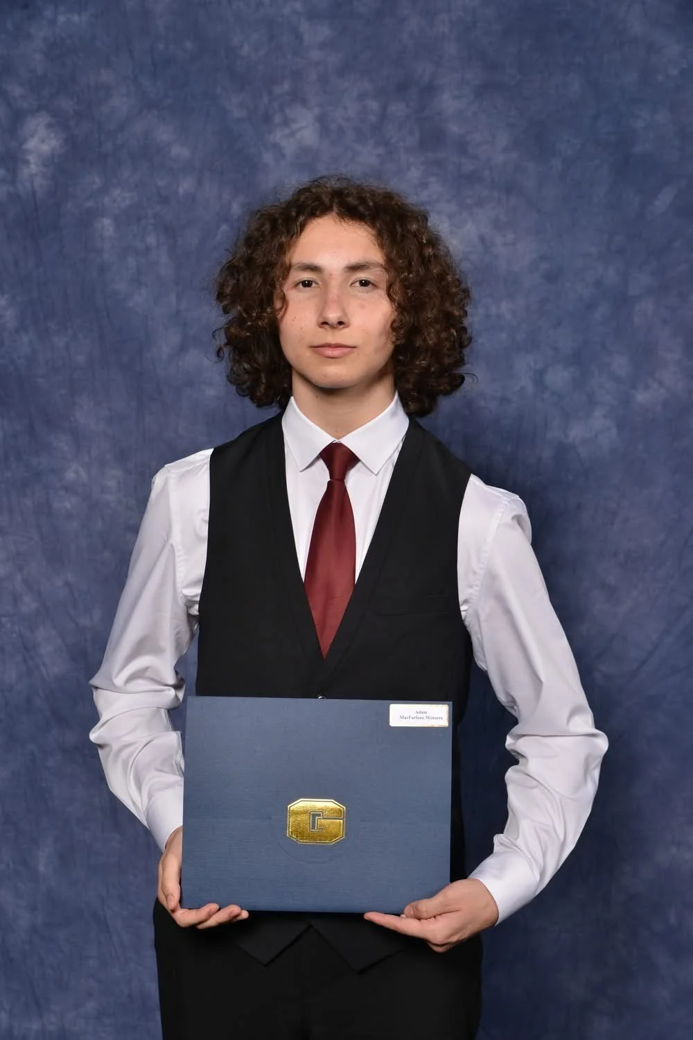 A young person with curly hair and light skin, dressed in a white shirt, black vest, and red tie, holding a blue diploma cover with a gold letter 'G' emblem, standing against a blue textured background.