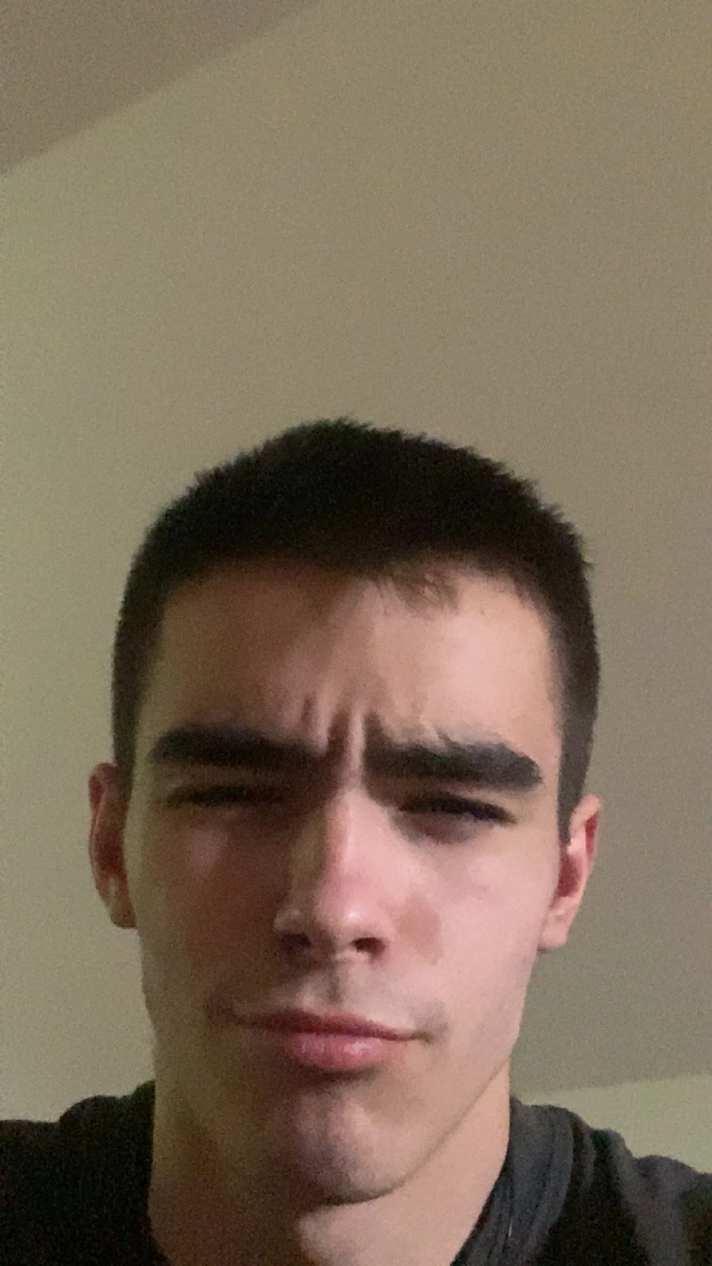 A young man with dark hair and thick eyebrows makes a frowning expression in a selfie against a neutral background.