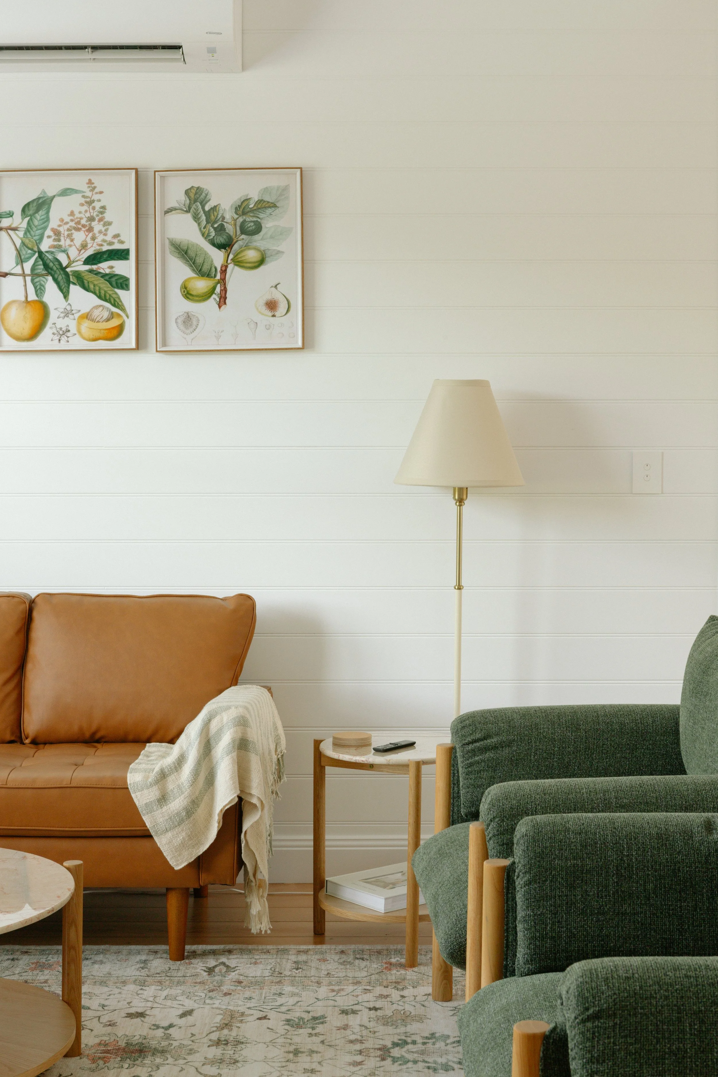 A living room with a brown leather sofa, a green armchair, a small wooden side table, a floor lamp with a beige shade, two framed botanical prints on a white wall, and a patterned rug.