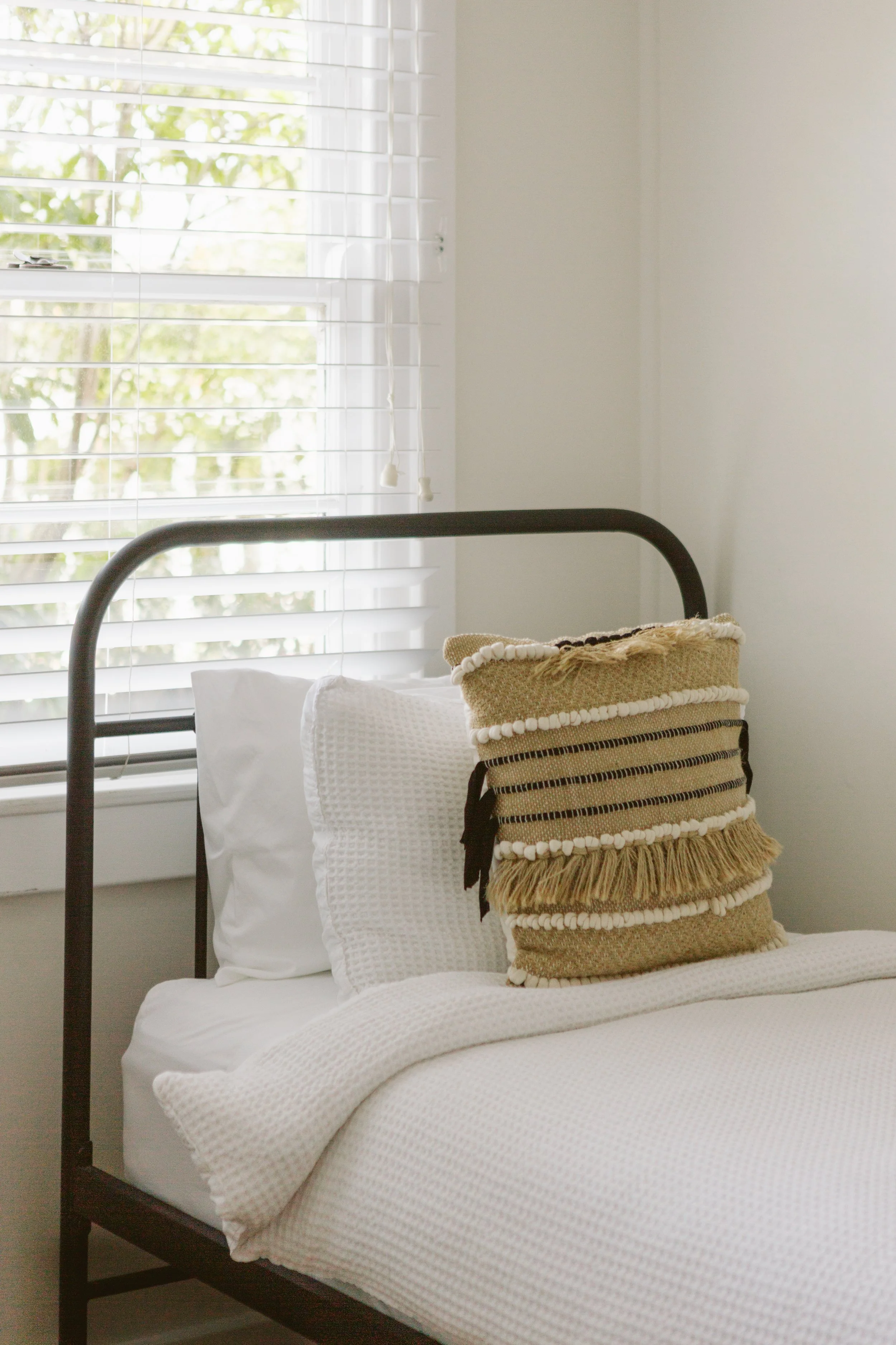A neatly made bed with white bedding and pillows, and a decorative pillow with tassels, next to a window with white blinds and greenery outside.