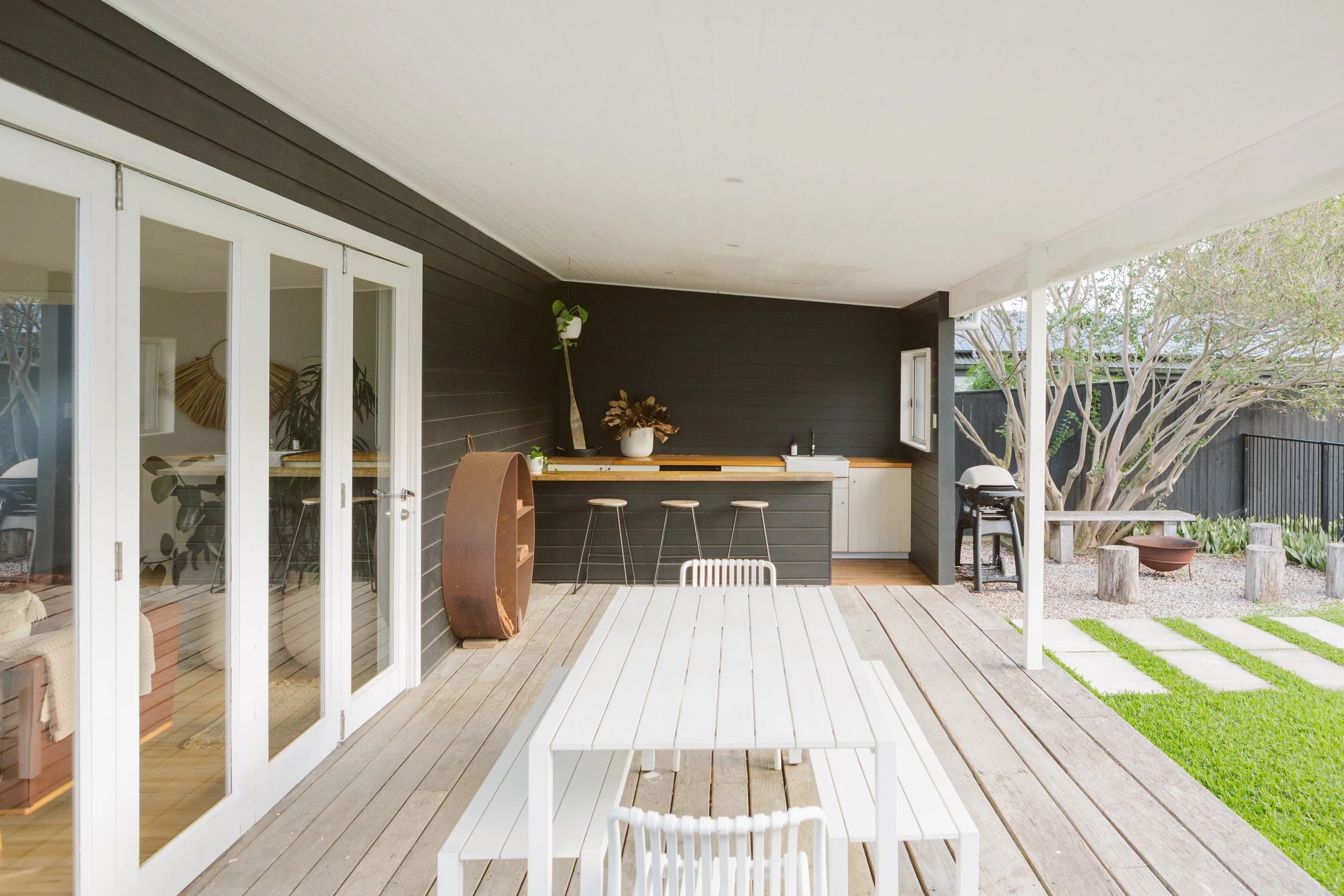 Outdoor covered patio with white wooden furniture, black wall, and a small kitchenette area with bar stools, potted plants, and a barbecue grill, adjacent to a grassy yard with stepping stones.