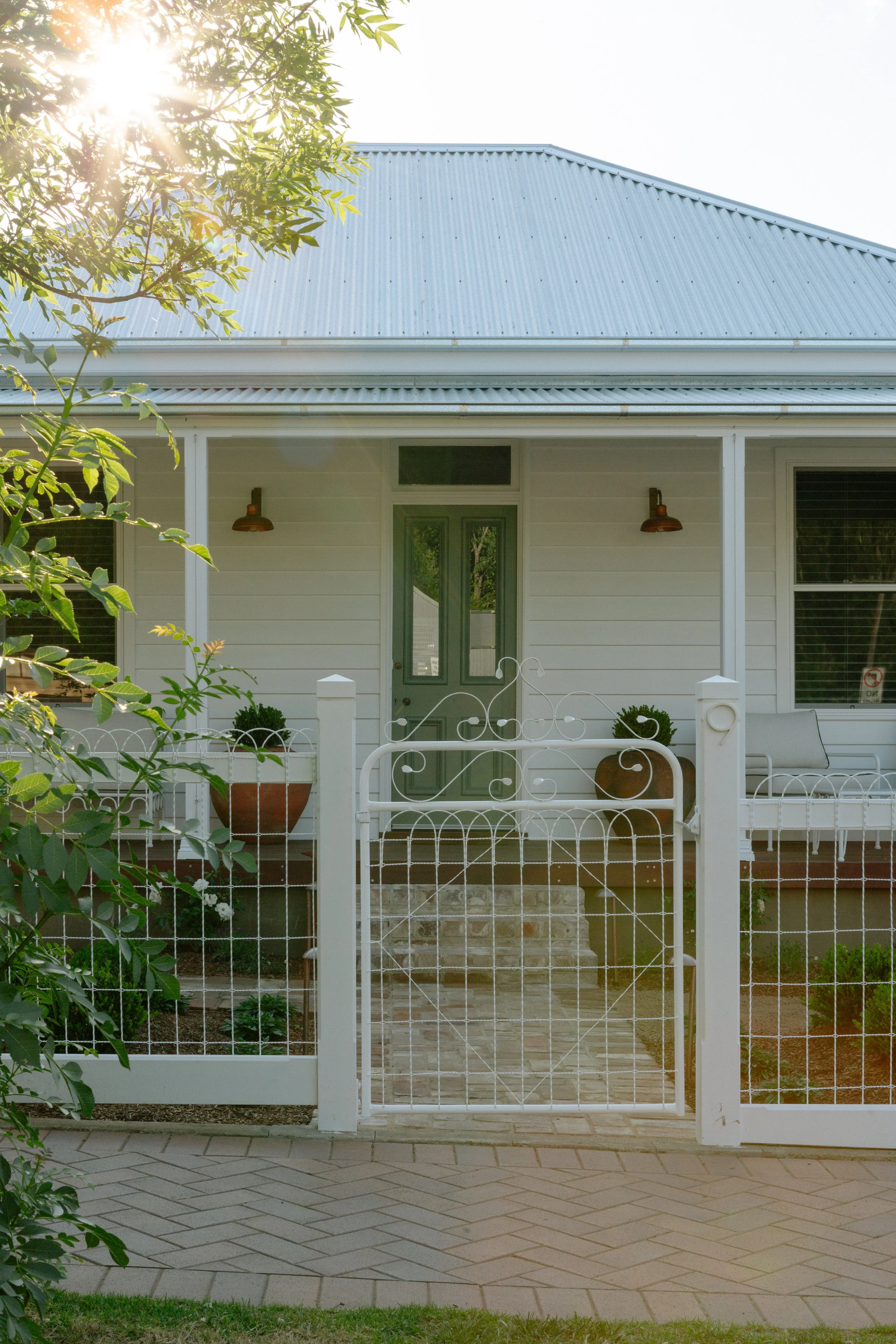 Front view of a white house with a light gray door, two wall-mounted lamps, a white fence with a gate, and potted plants in the front yard, captured in bright sunlight.