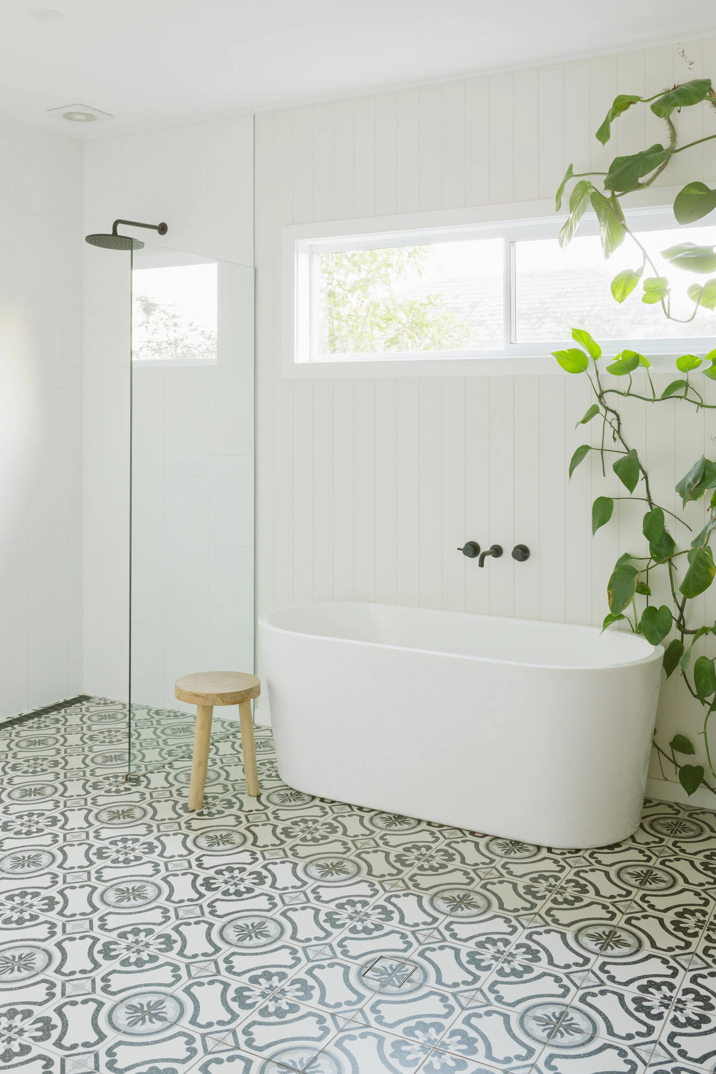 A clean, modern bathroom featuring a freestanding white bathtub, a small wooden stool, a shower area with a black showerhead, a large window, white paneled walls, and patterned black and white floor tiles with a green plant growing next to the bathtu