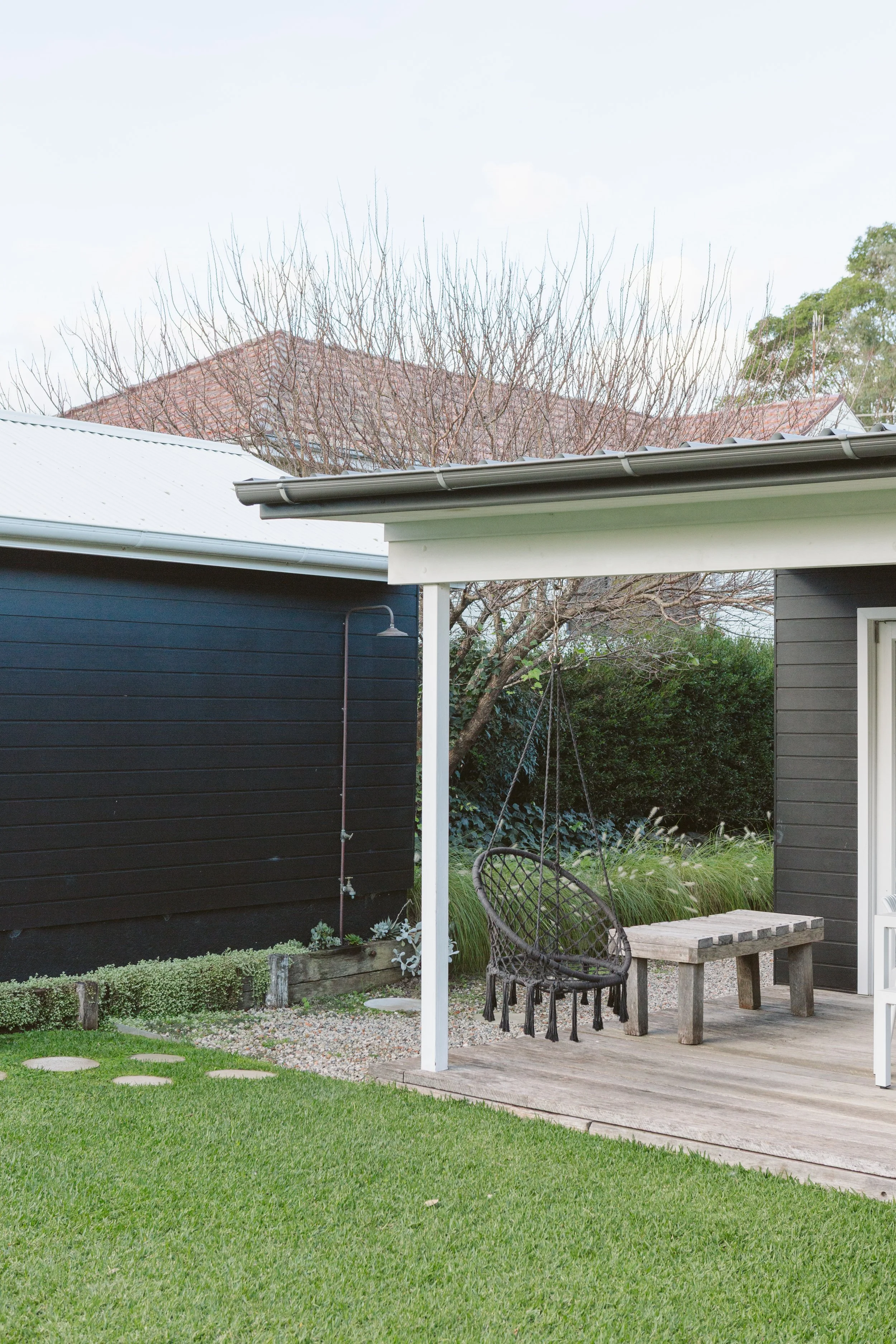 A backyard patio with a hanging chair, a wooden bench, part of a house, and a lawn with stepping stones.