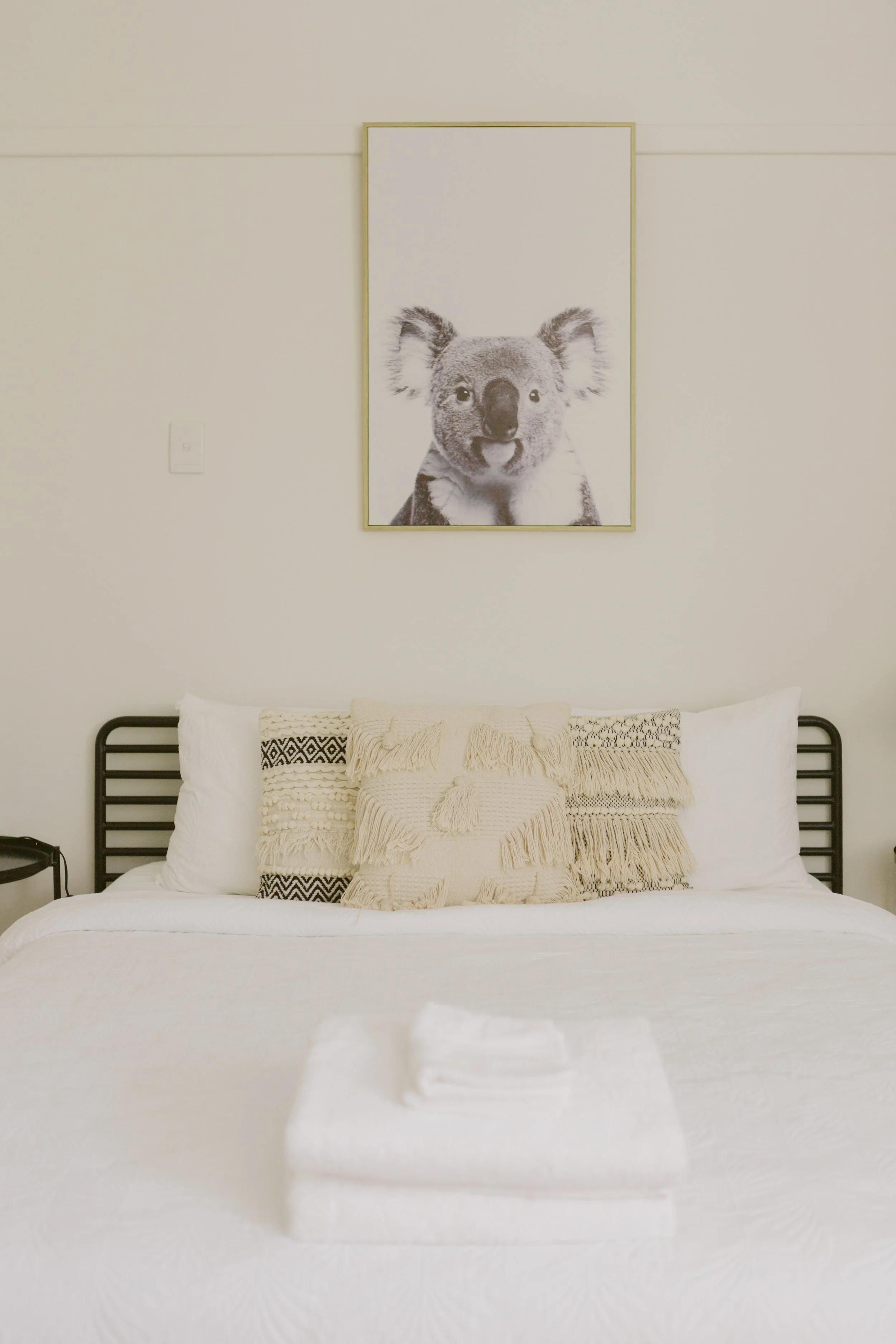 A neatly made bed with white bedding and decorative pillows, topped with folded white towels, in front of a simple wall with a framed black and white portrait of a koala.
