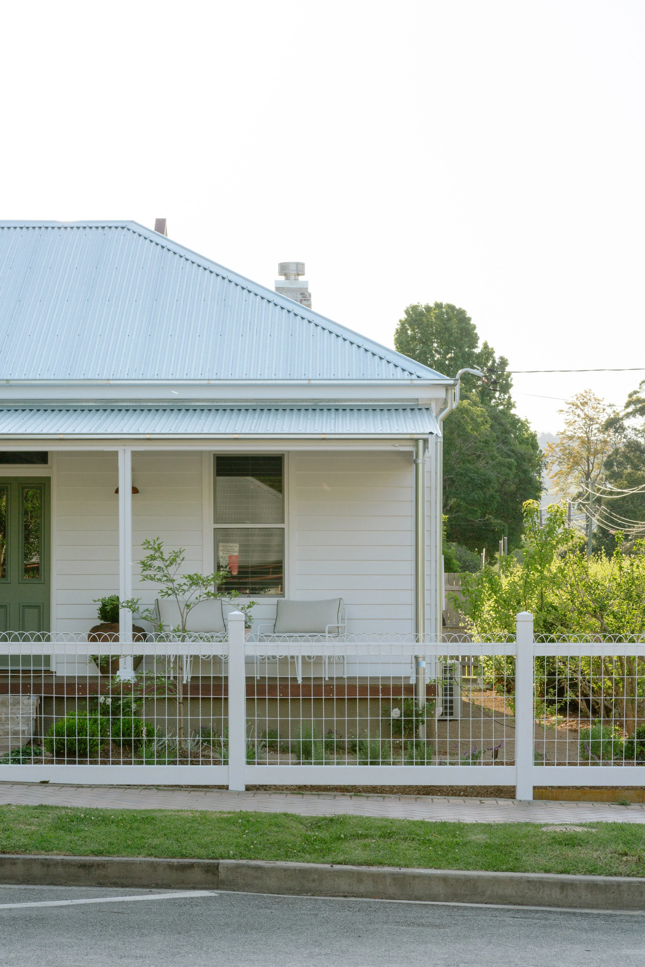 Front yard view of a white house with a green door, sitting on a small porch with two bench chairs and potted plants, enclosed by a white picket fence, with a garden and trees in the background.