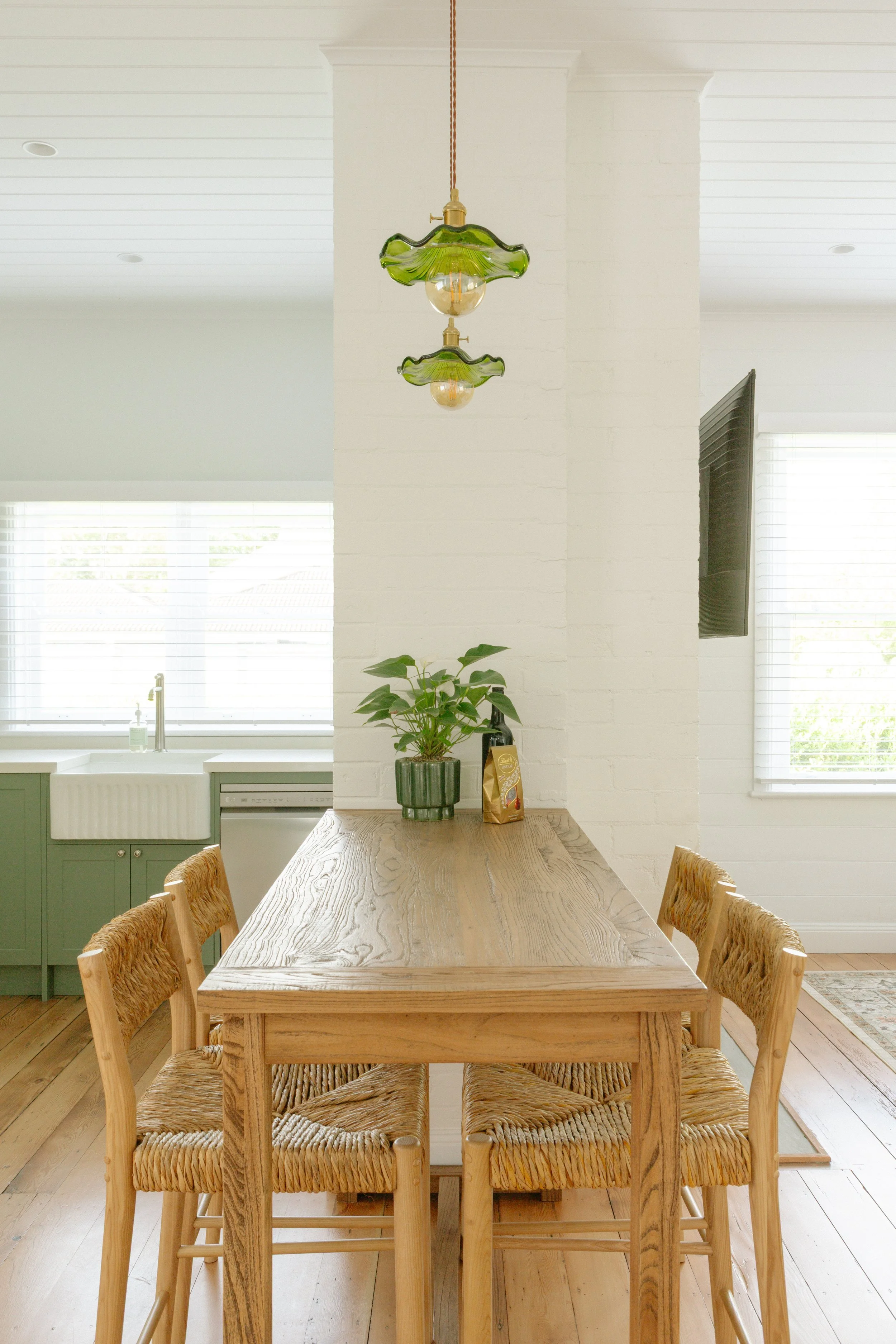 A dining area with a wooden table, four wooden chairs with woven seats, a green potted plant, a bag of coffee, hanging green glass pendant lights, and a wall-mounted TV in a bright room with large windows and white walls.