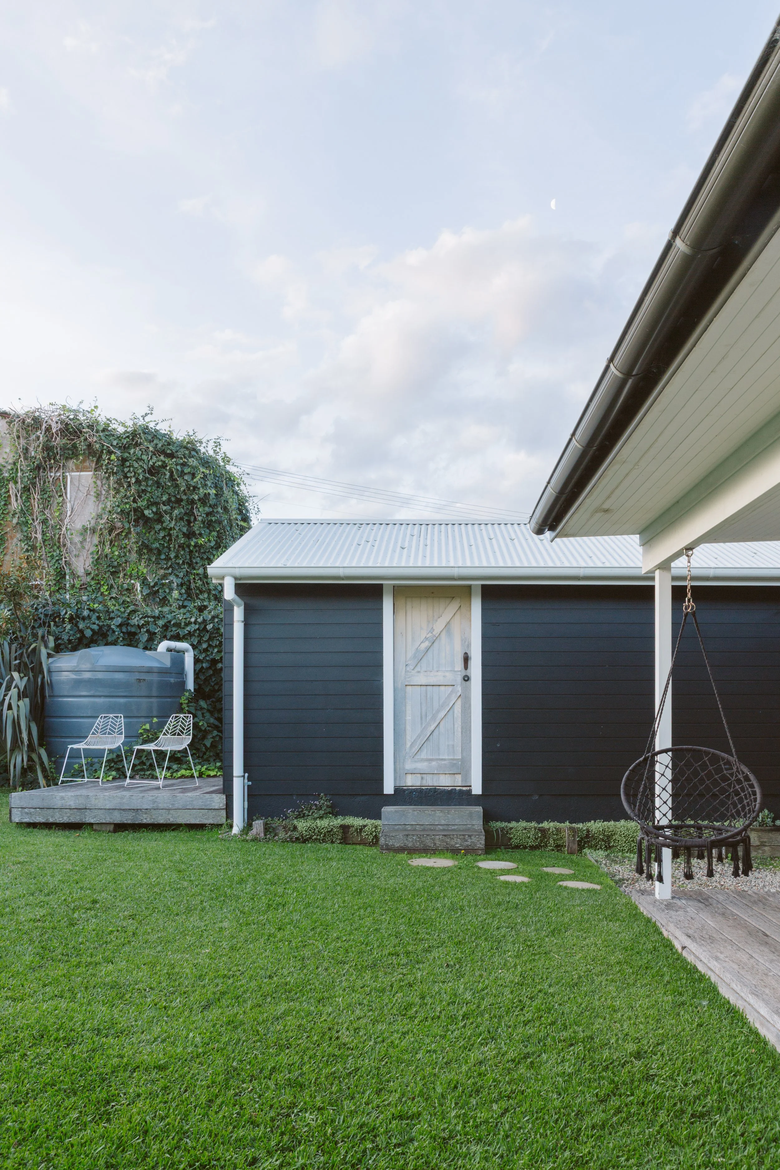 Backyard with green grass, small black building with white door, white two chairs, water tank, and hanging chair, under cloudy sky.