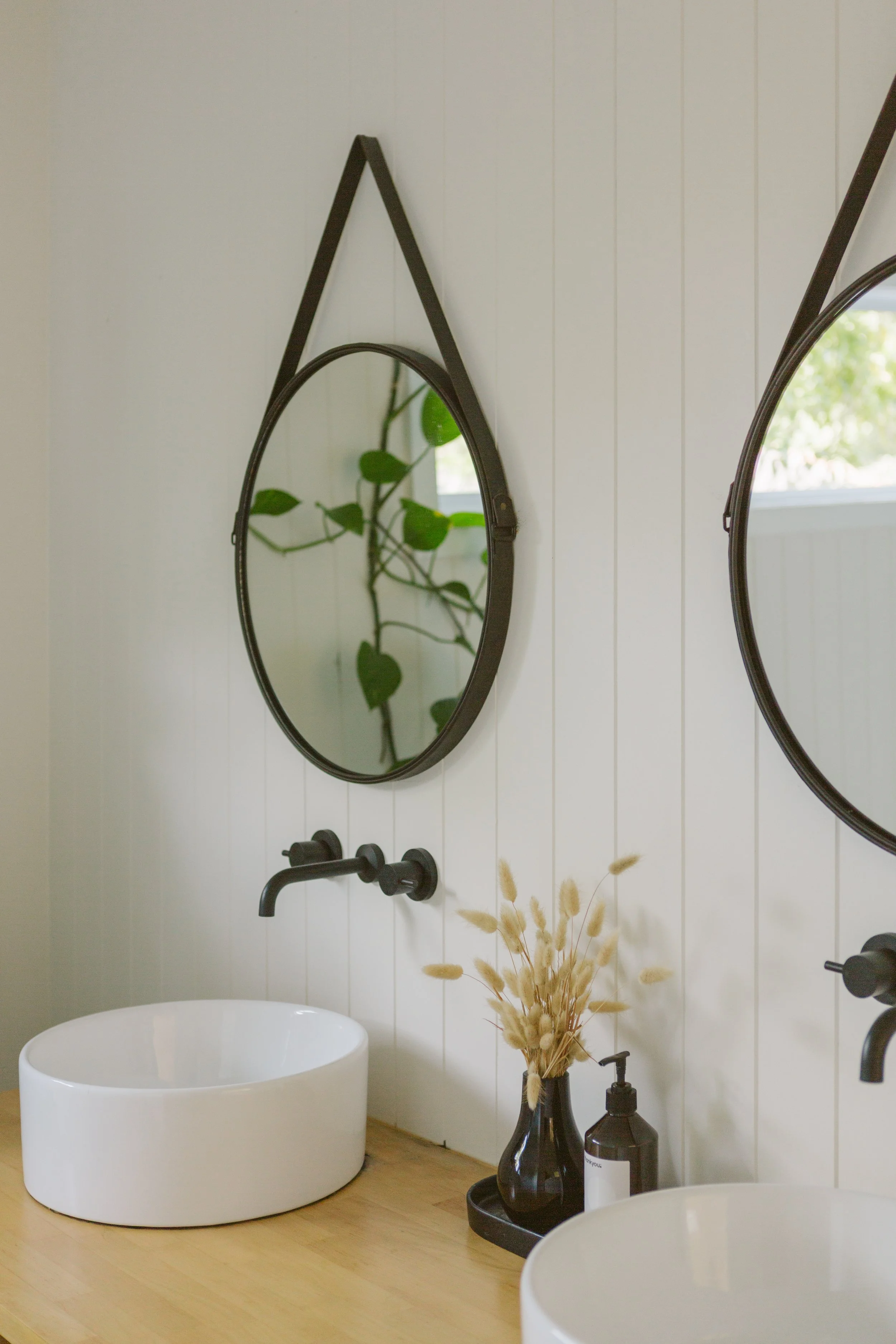 Modern bathroom with two vessel sinks on a wooden countertop, black wall-mounted faucets, and two round mirrors with black frames hanging on a white paneled wall. A black vase with dried beige grass and a black soap dispenser are placed between the s
