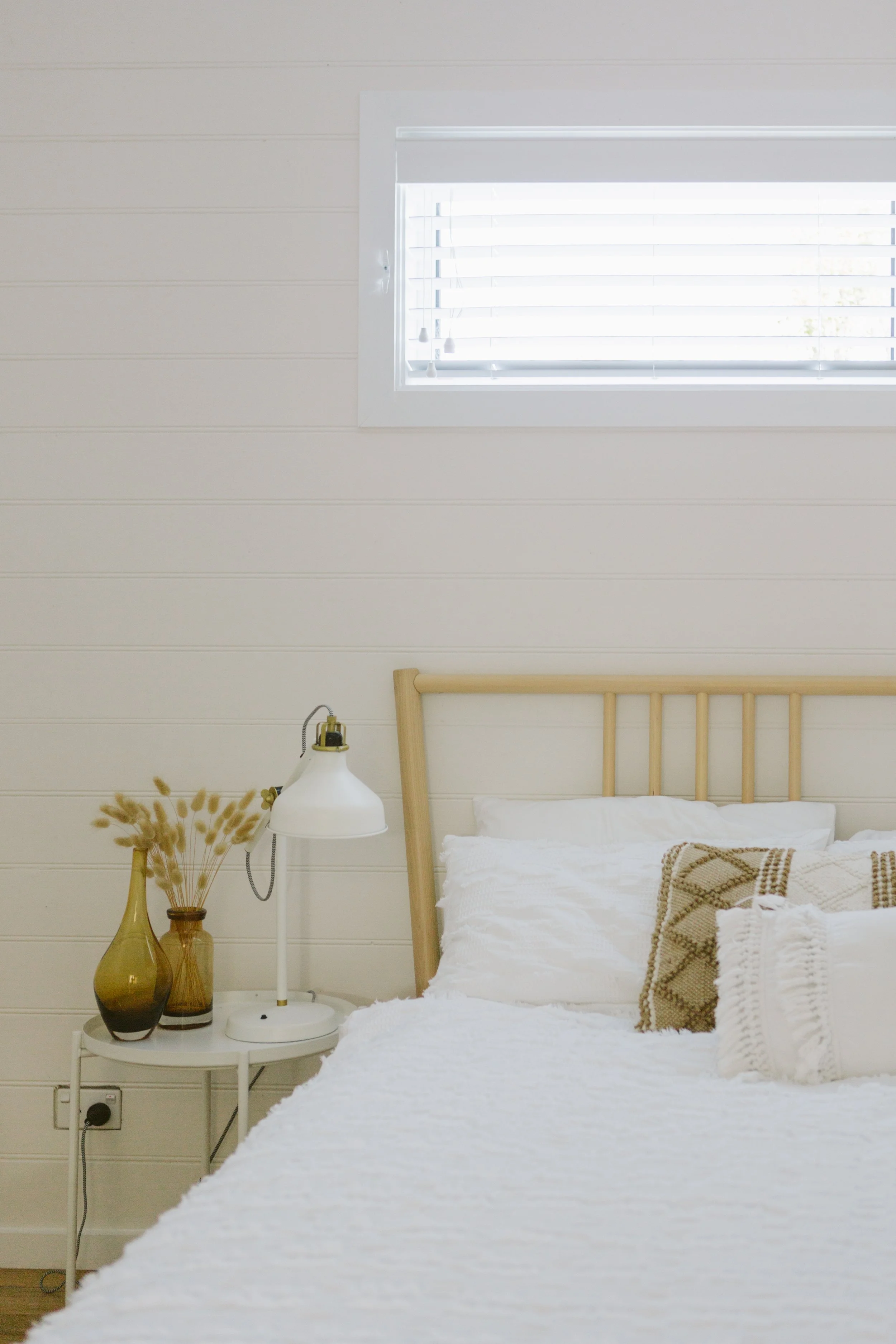 A cozy bedroom with cream-colored walls, a white bedspread, and decorative pillows. A small white side table holds two brown vases with dried flowers and a white lamp. There is a window with white shutters above the bed.